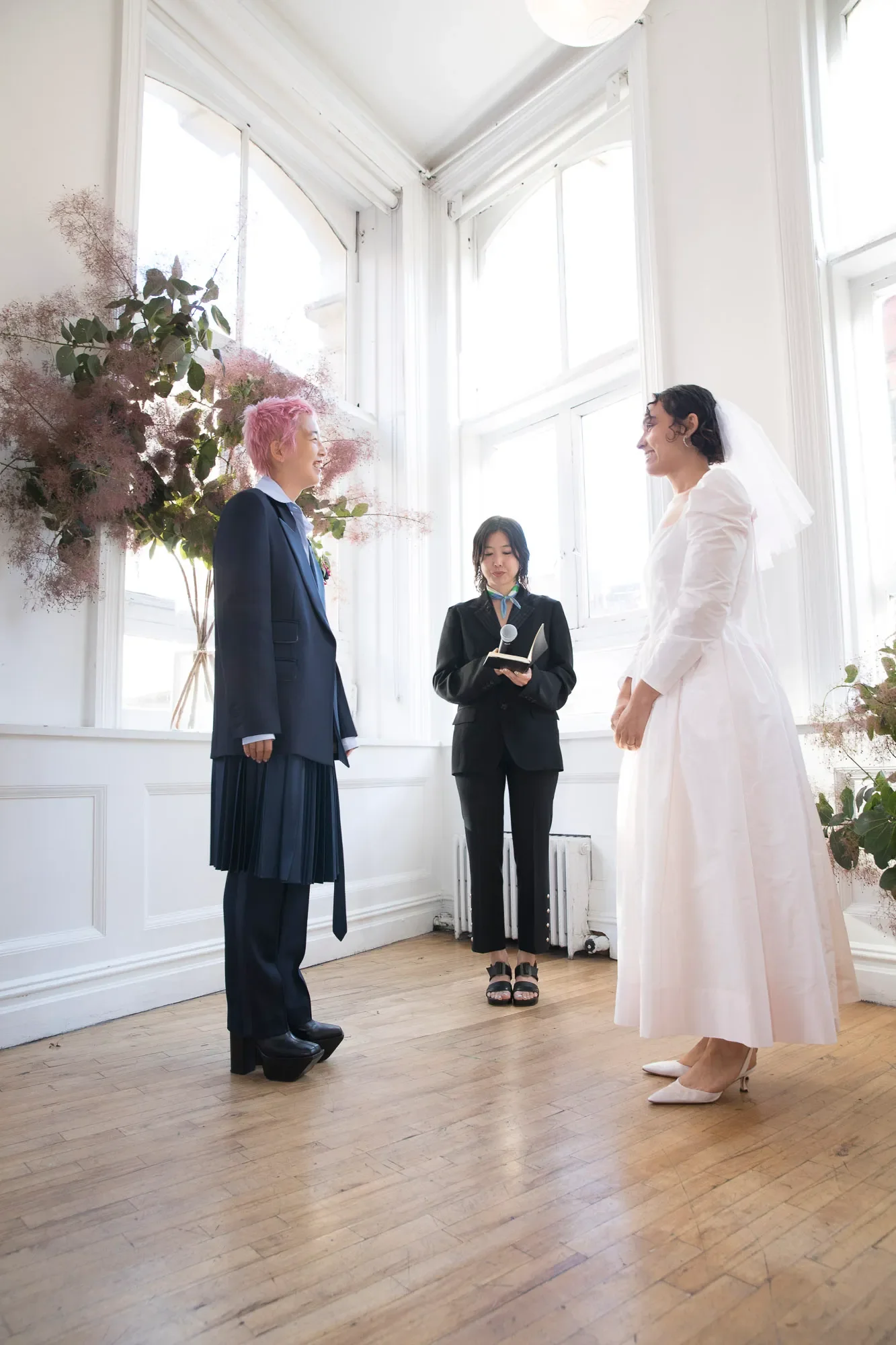 Two people stand in front of an officiant in a bright white room with large windows. The person on the left is wearing a suit and has cropped pink hair. The person on the right is wearing a long sleeved white gown and has a dark bob.