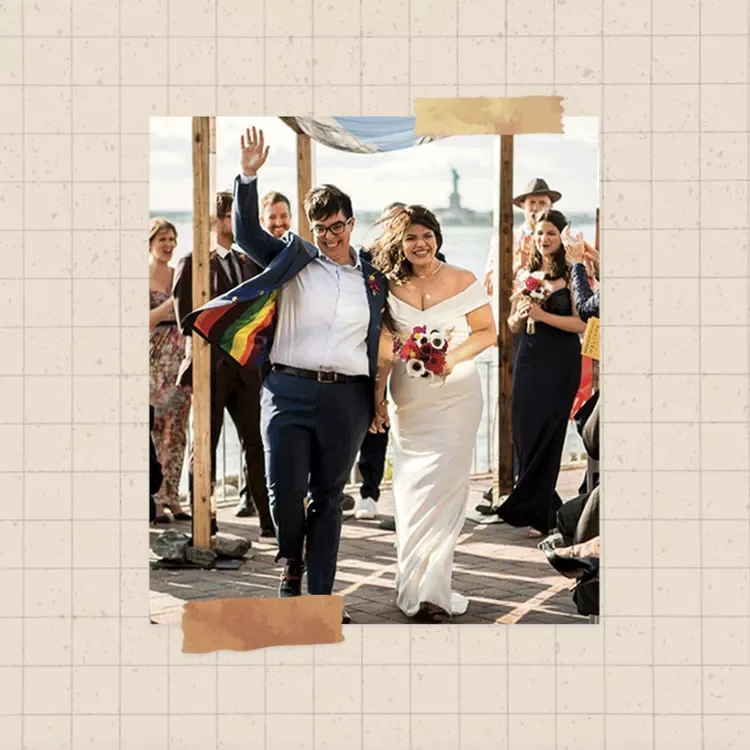 A couple walks down the aisle at an outdoor wedding with their loved ones cheering behind them. One of the couple is wearing a dark suit with a rainbow lining, the other is wearing an off the shoulder white gown.