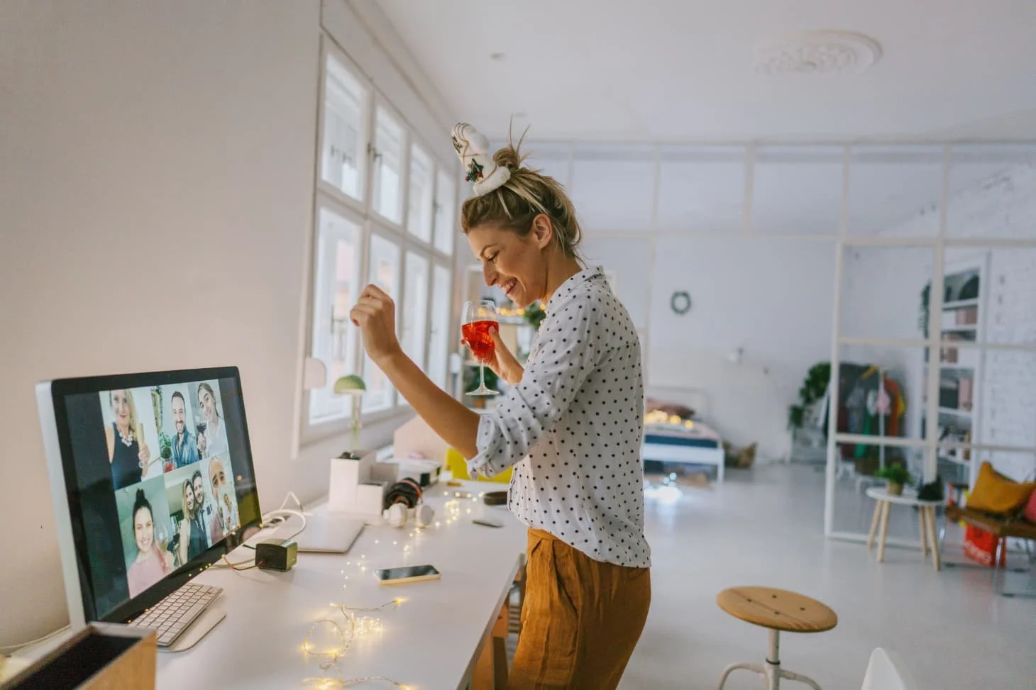 A woman wearing a party hat stands in front of a computer screen and cheers. On the screen is shown a virtual meeting software with multiple people in the call.
