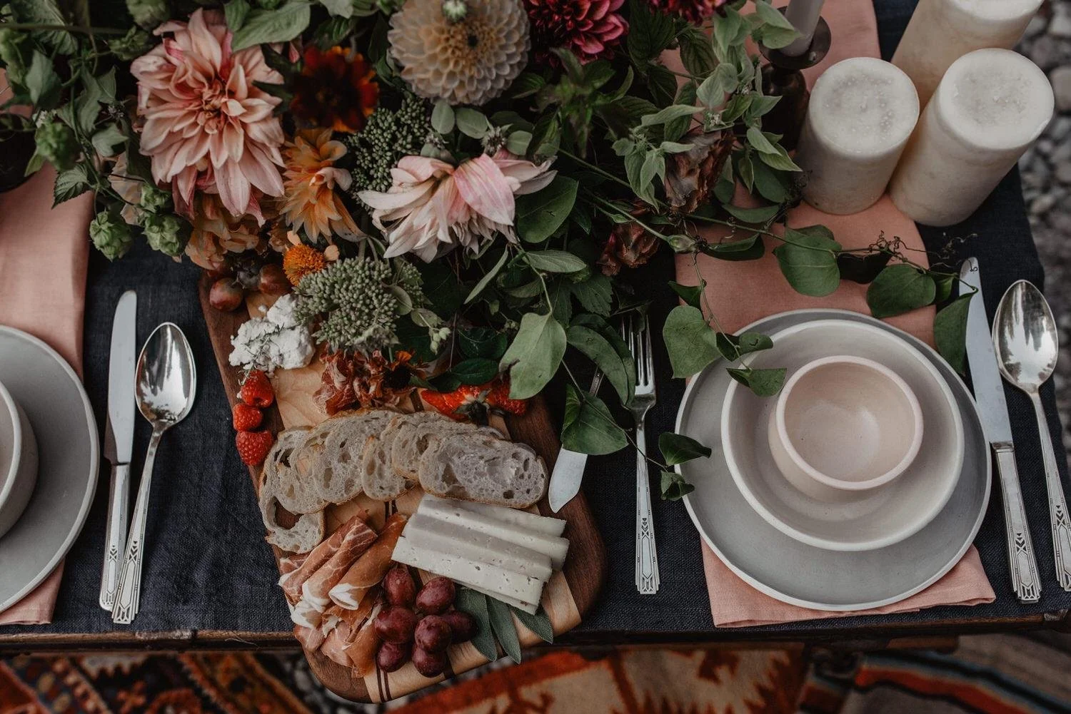 Moody image with a table setting. Candles, muted greenery, and dusky blue napkins are shown. On the table is a charcuterie and bread display.