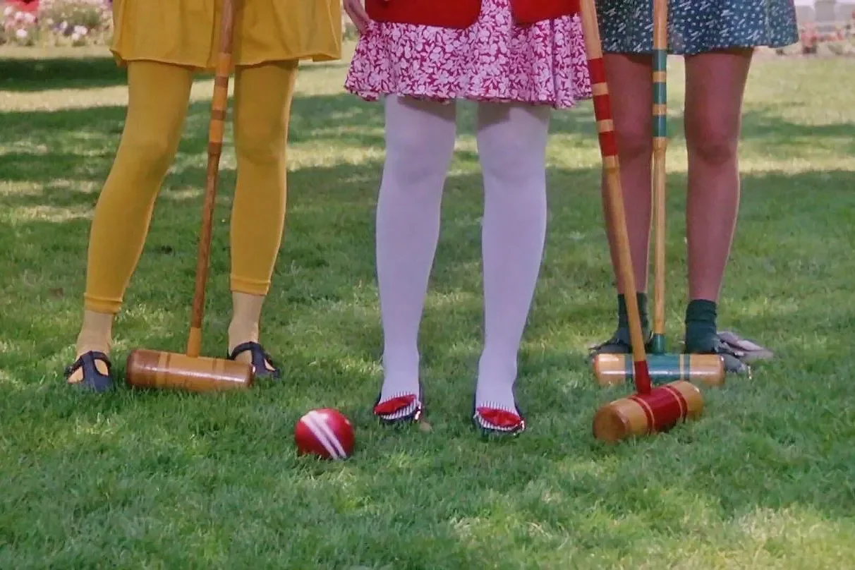 Three people stand in festive outfits on a lawn wearing yellow, purple, and blue outfits from left to right. All are pictured with croquet mallets and balls.