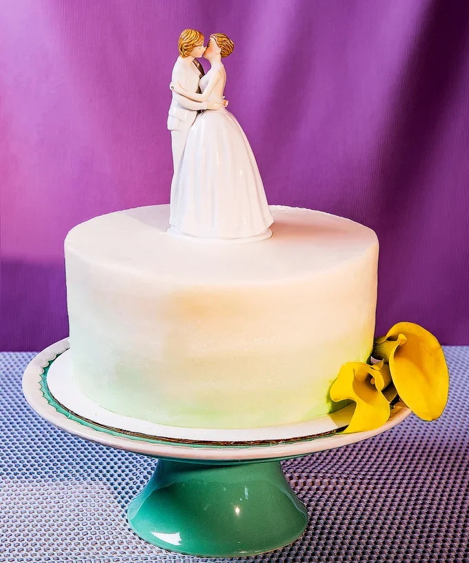 A white wedding cake with a topper of two LGBTQ partners is shown. The cake is on a green cake stand and shown against a purple backdrop.