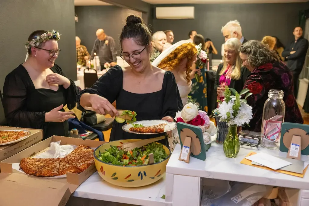 Brunette woman with glasses shown serving herself food from a yellow bowl. Behind her is a room full of people gathered.