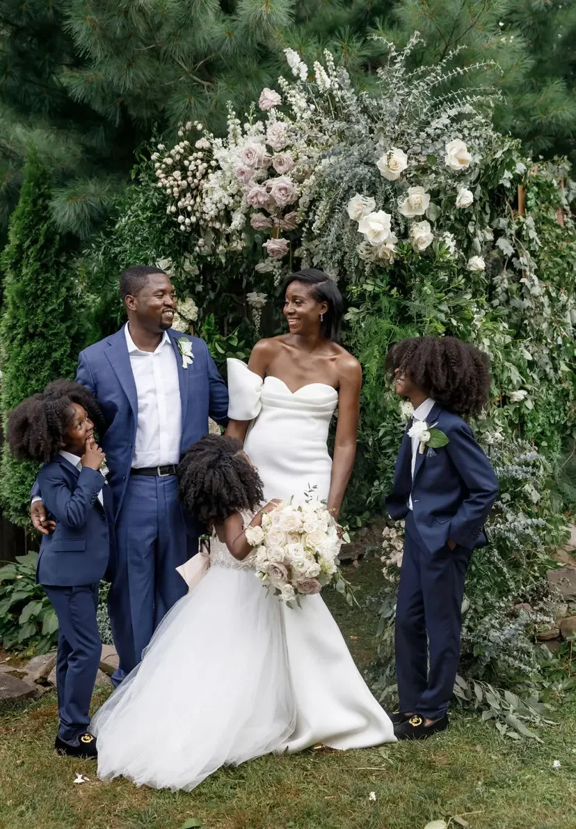 A couple is pictured at their wedding with three children. Two children and the groom wear dark suits, the third child and the bride wear white gowns. All are smiling.