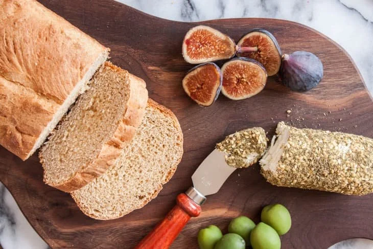 A charcuterie platter with sliced wheat bread, figs, herbed goat cheese, and green olives. Also on the platter is a red-handled cheese knife.