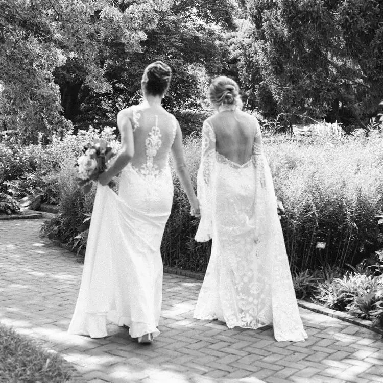 Two brides face away from the camera, both holding bouquets. The background is a garden path.