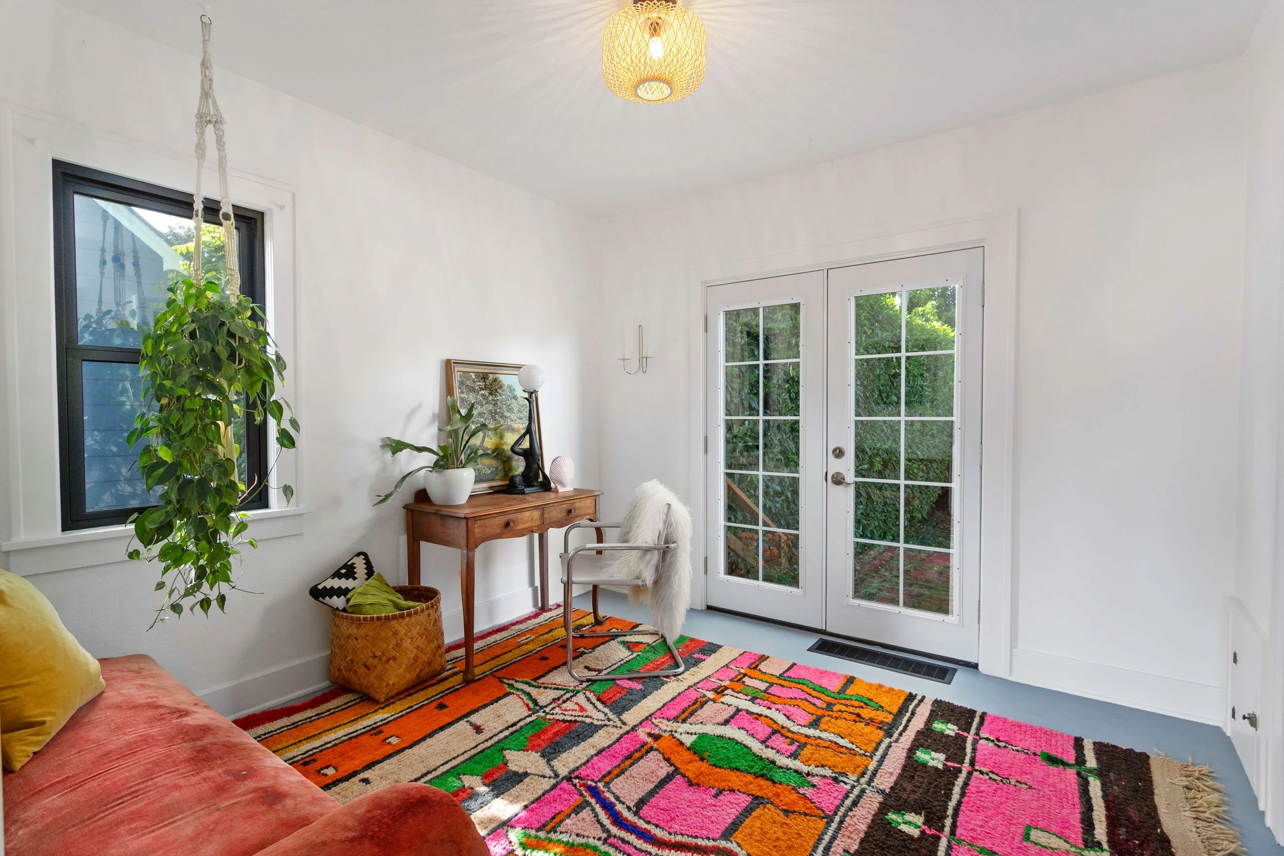 Bright living room with white walls, colorful rug, orange velvet sofa, green hanging plant, wooden side table with decorative items, white arm chair with fur throw, glass double doors leading to garden, window with greenery outside, ceiling light fixture.