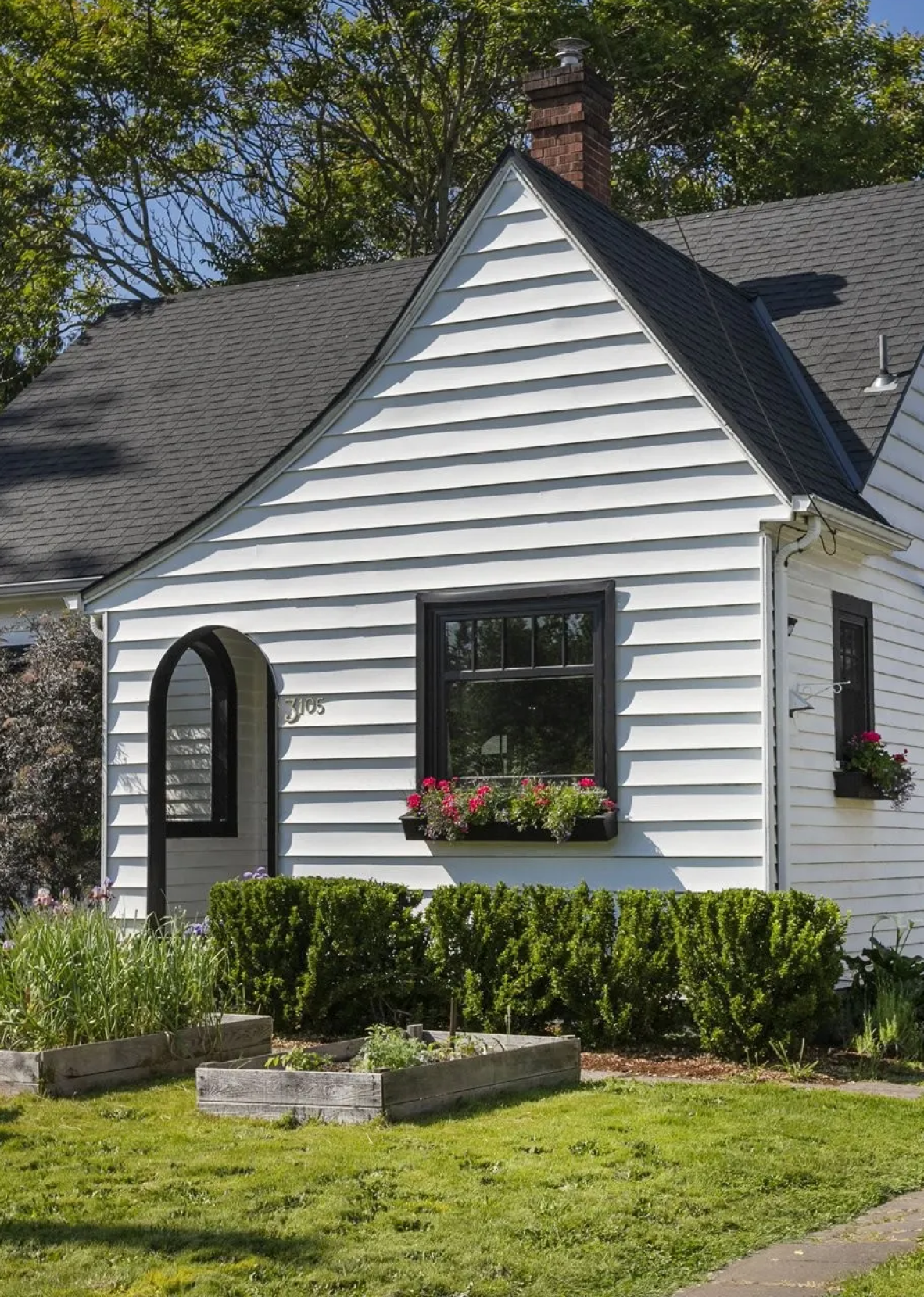A white house with black window frames, a black arch doorway, and a gray shingled roof amidst green trees with flower boxes on the windows and a manicured garden in the front yard.