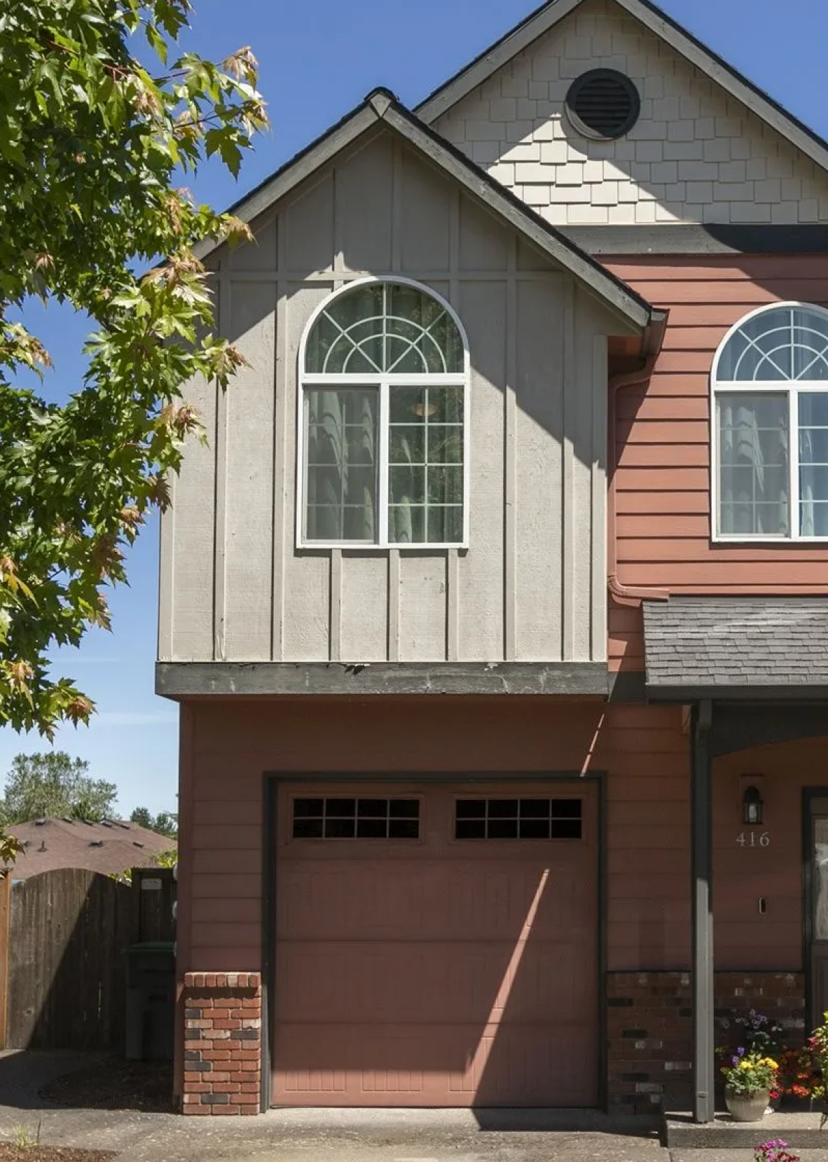 Close-up exterior of a two-story house with a garage, red and beige siding, arched windows, and a tree partially visible on the left side.
