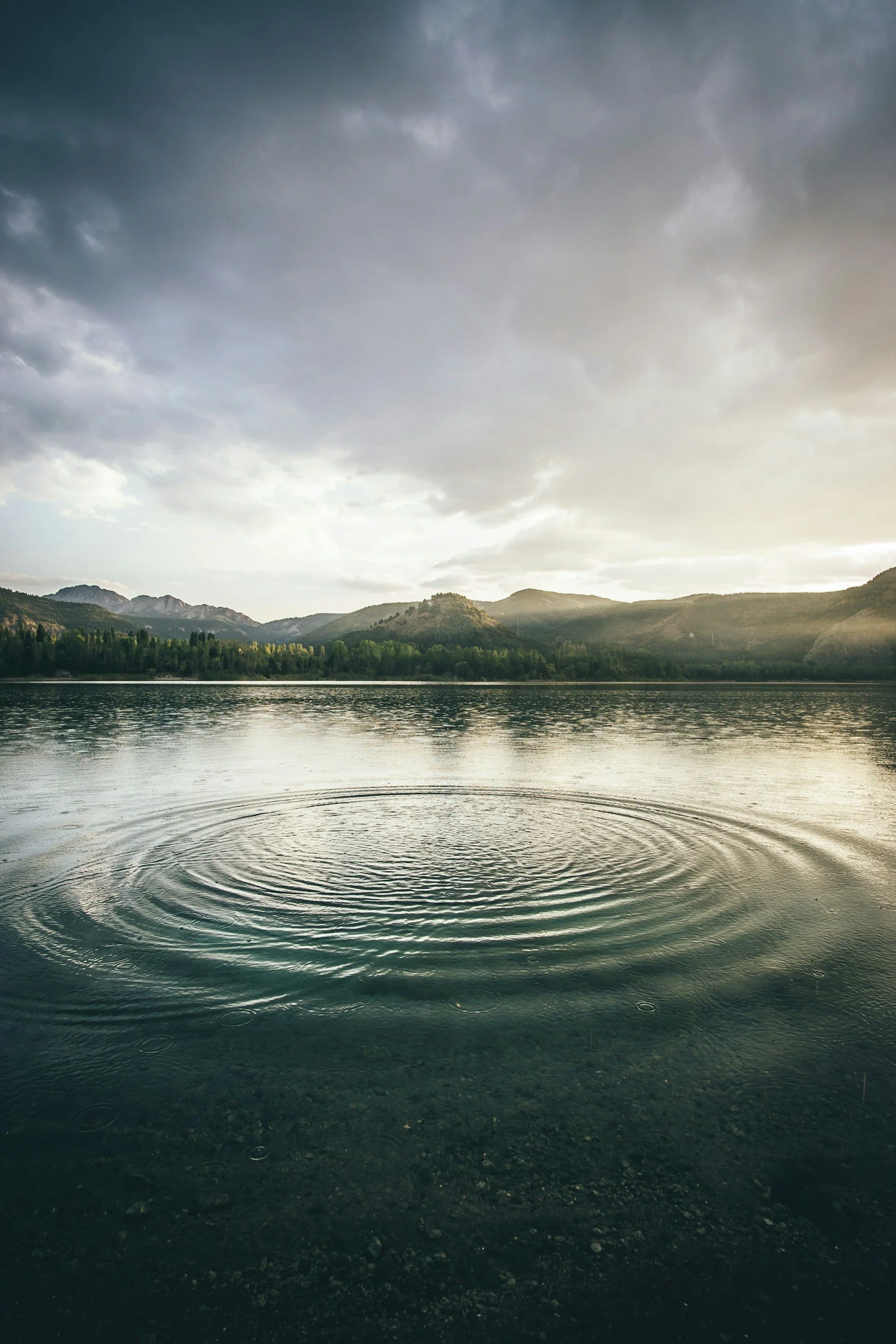 A tranquil lake with gentle ripples, surrounded by green mountains under a cloudy sky at sunset.