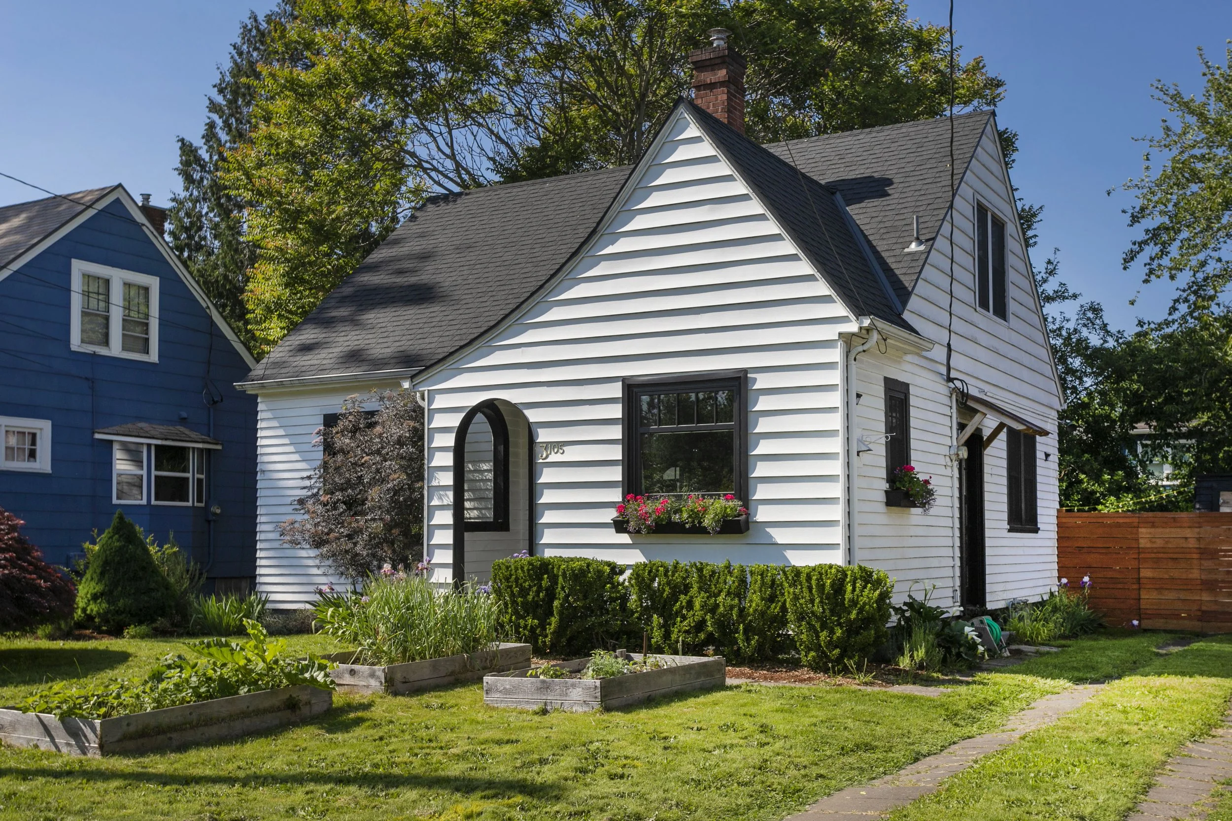 White two-story house with black window frames and a chimney, surrounded by a green lawn, gardens, and trees under a blue sky.