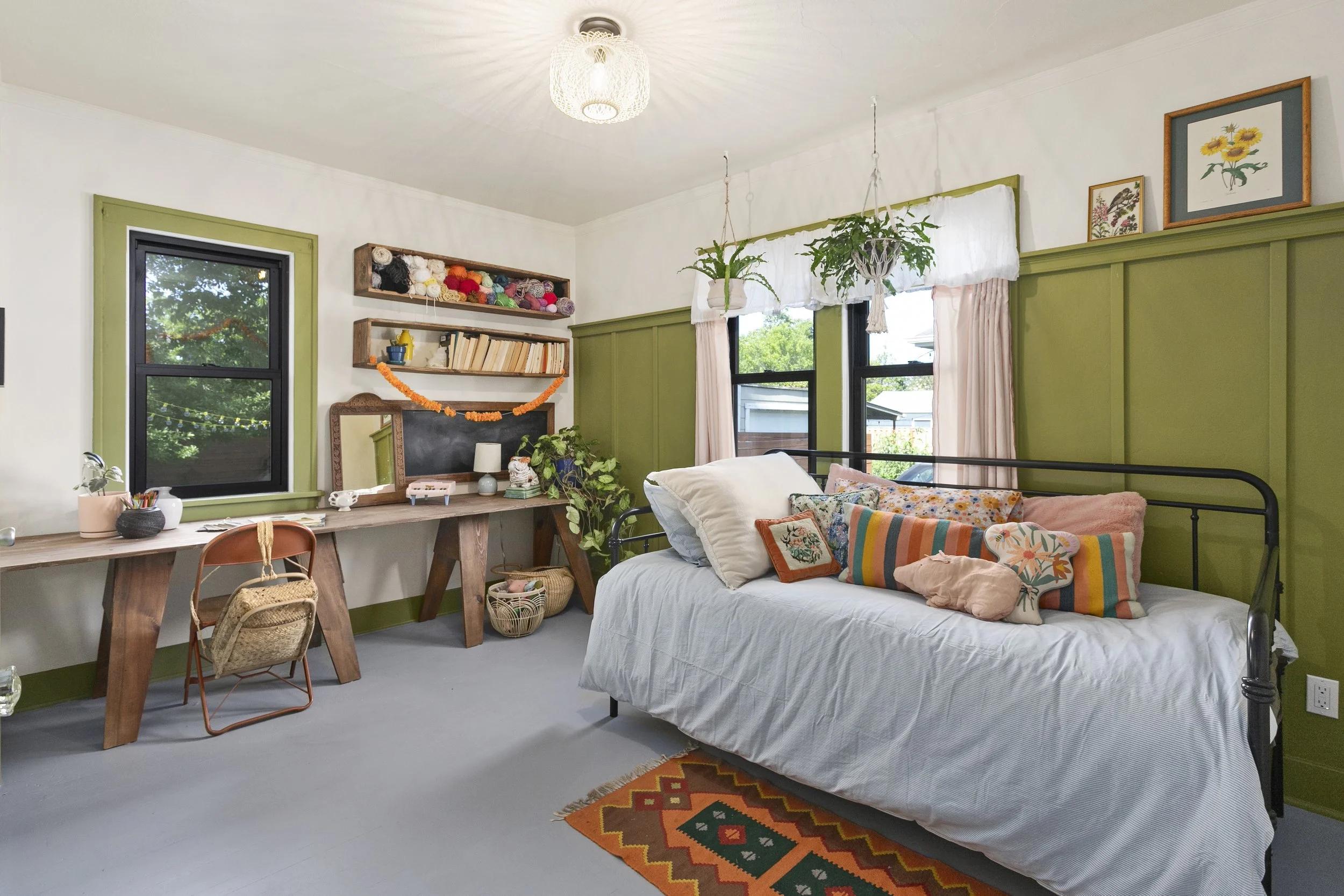 A cozy bedroom with a black metal bed frame, white bedding, and colorful throw pillows. The room has green wainscoting, windows with pink curtains, and hanging plants. There is a wooden desk with a mirror, books, and decor, along with a chair and a small rug featuring geometric patterns.