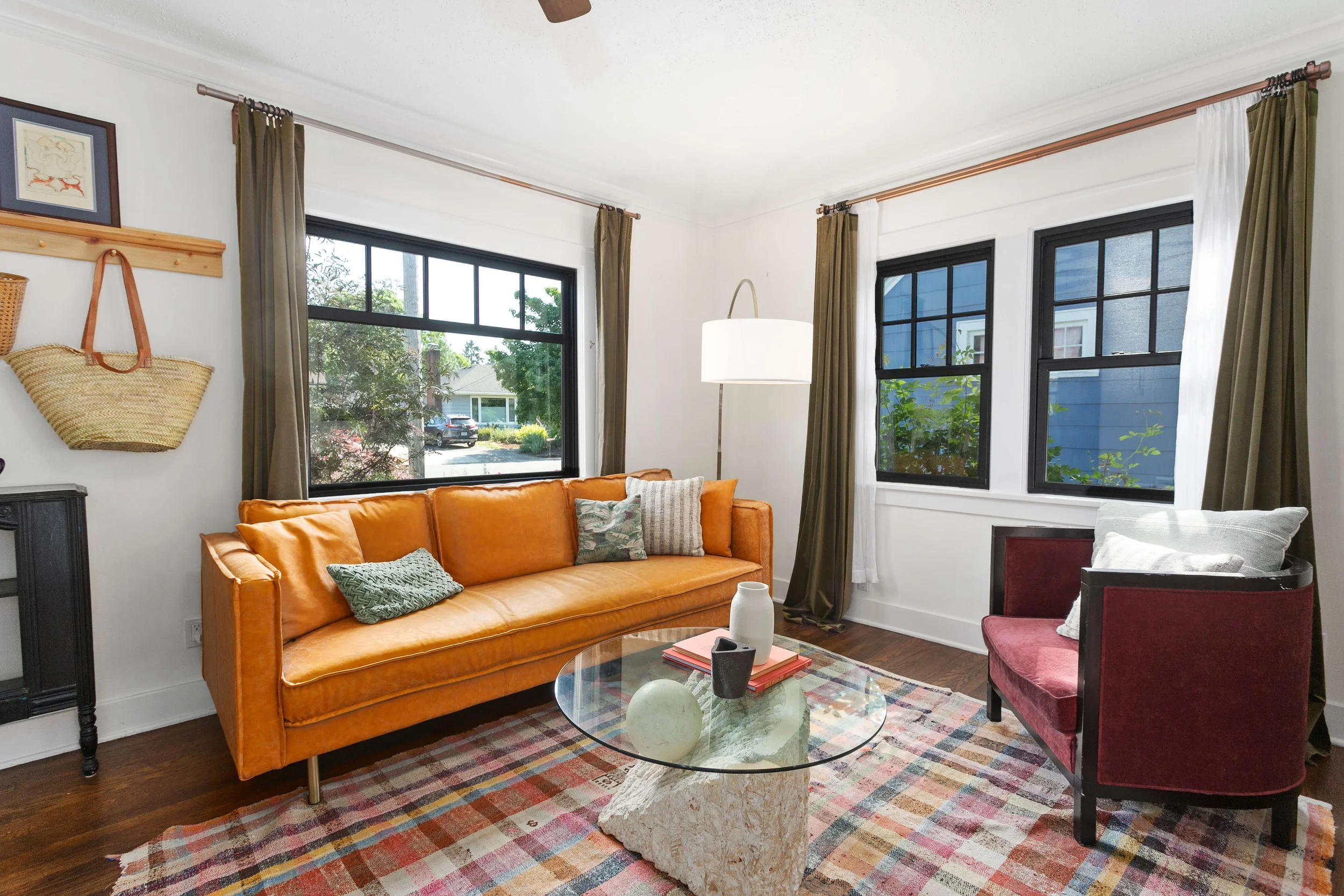 Living room with large windows, tan leather sofa, red armchair, glass coffee table with decorative items, colorful rug, and curtains.