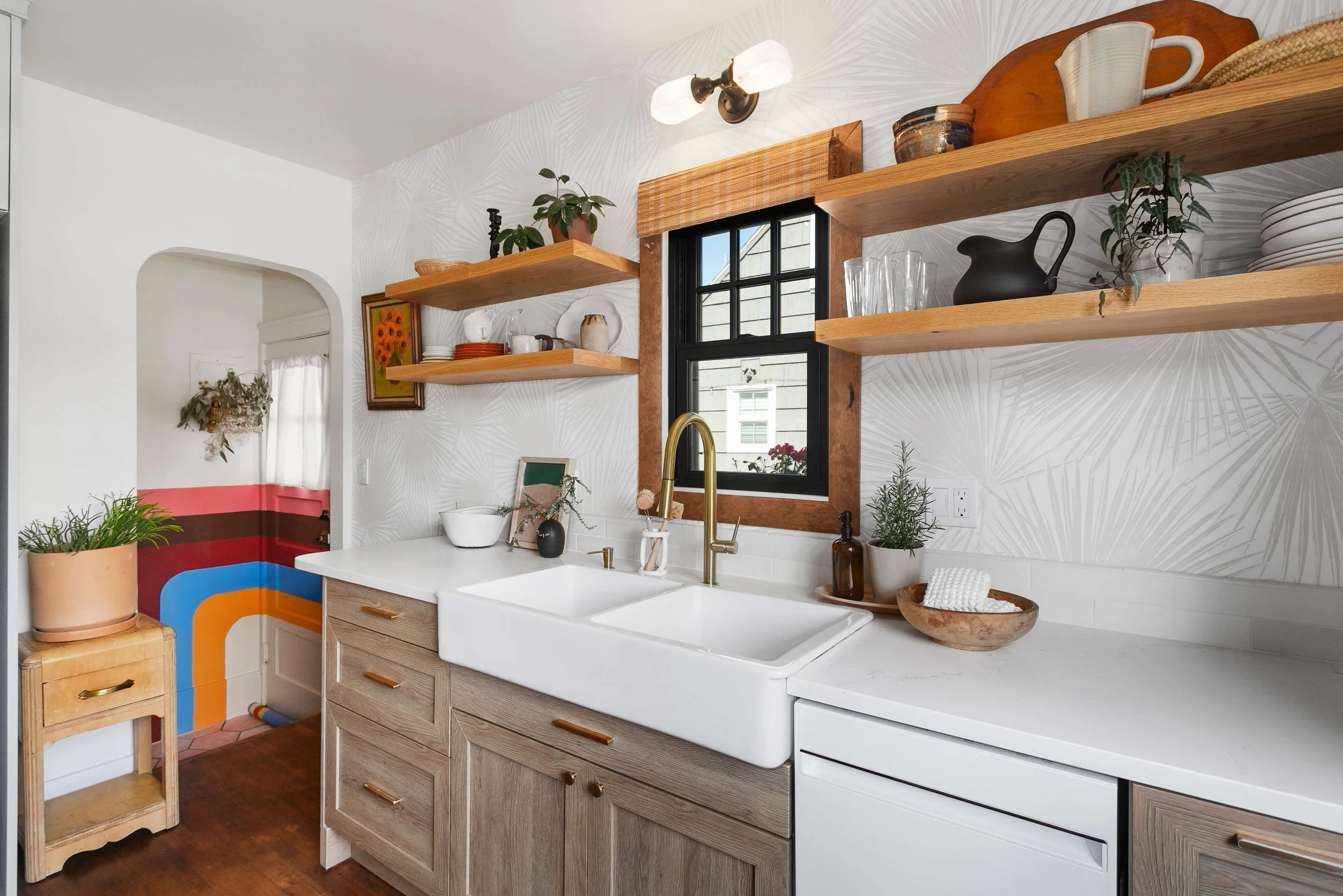 Kitchen with white farmhouse sink, wooden cabinets, open wooden shelves with dishes and plants, window with wooden trim, and a wall with patterned wallpaper.