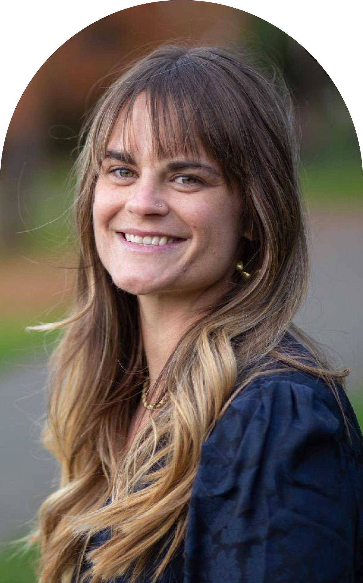 A woman with long, wavy hair smiling outdoors, wearing a dark blue top and jewelry.