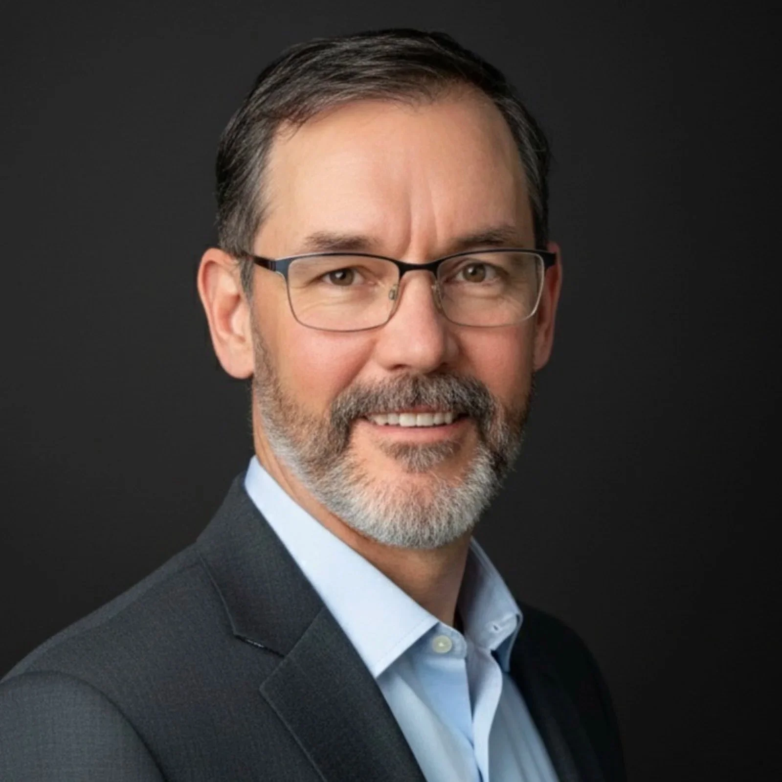 A professionally dressed middle-aged man with glasses, short dark hair, and a beard, smiling against a dark background.