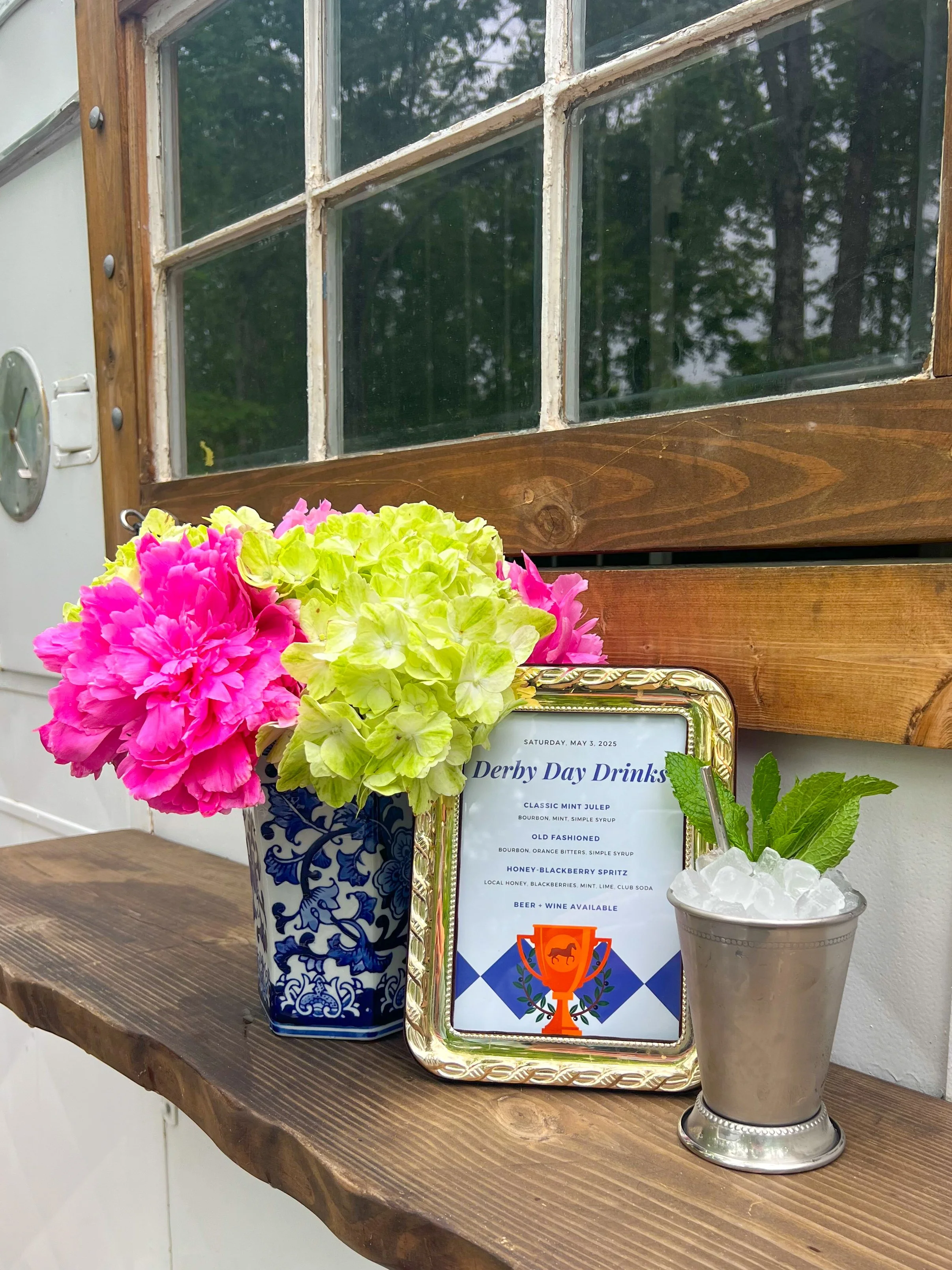 Flower arrangement and a framed menu for Derby Day Drinks on a wooden shelf against a wall with a window.