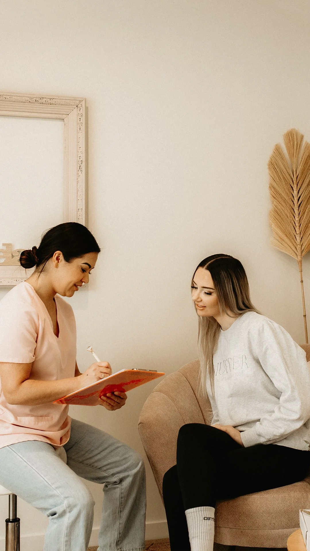 A woman in a pink shirt taking notes while talking to another woman sitting on a beige sofa in a cozy room with a large leaf decor on the wall.