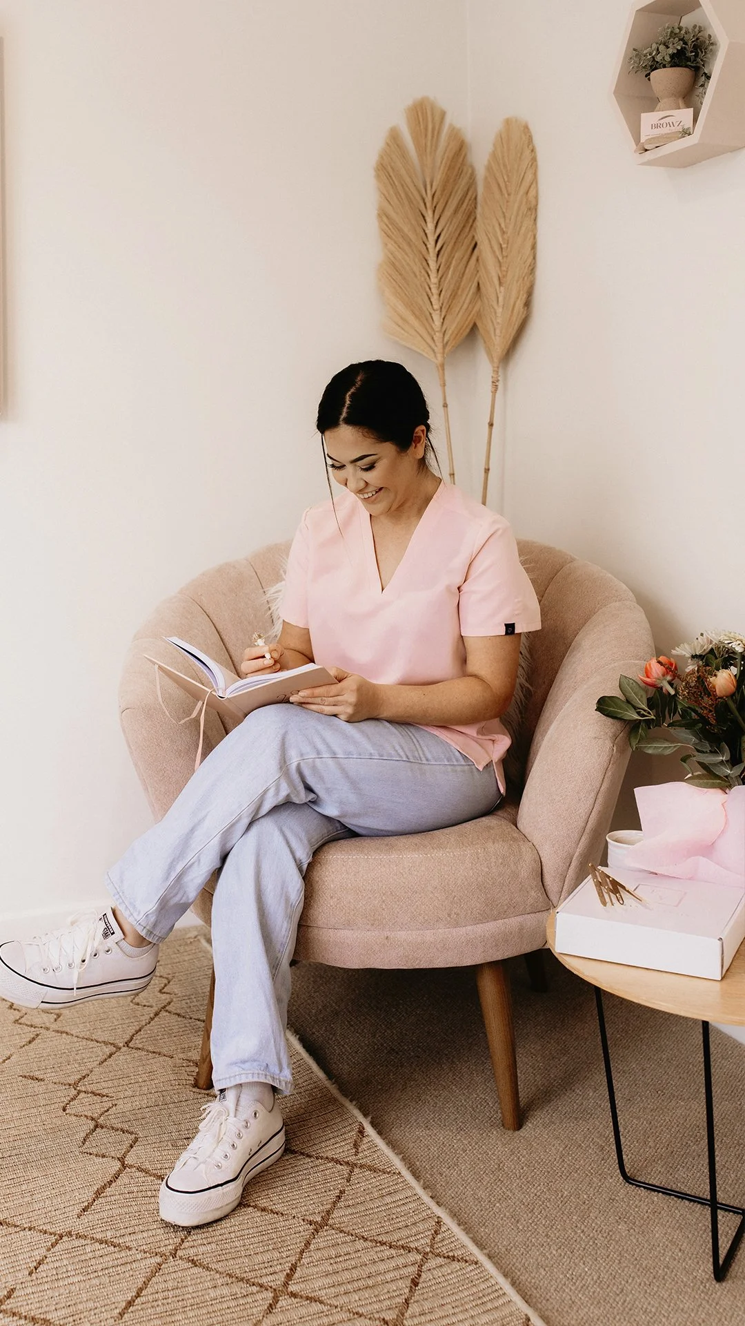 A woman with dark hair, wearing a light pink top and light blue jeans, sitting on a beige armchair. She is smiling and writing in a notebook. Behind her, there are two dried palm leaves in a decorative arrangement on the wall. To her right, there's a round side table with a bouquet of flowers in a gift box and some pink tissue paper on it.