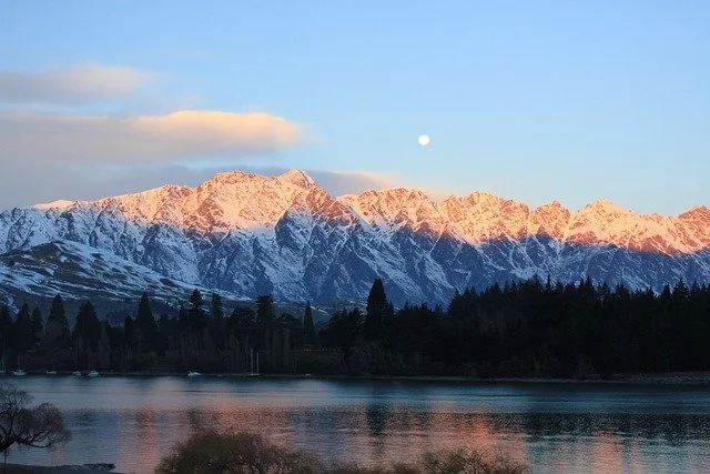Snow-capped mountains at sunrise or sunset with a lake in the foreground and a clear sky with a visible moon.