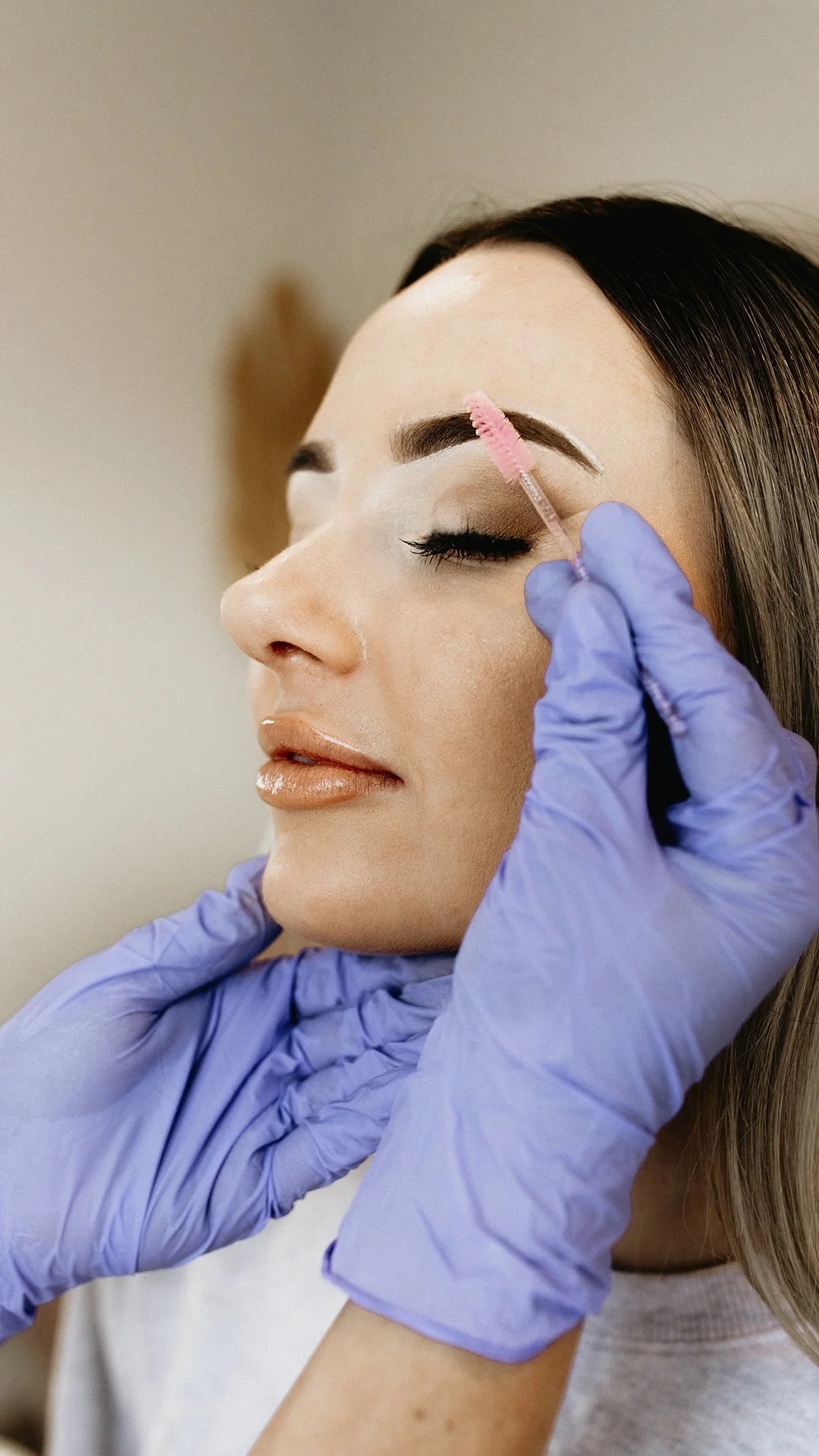 A woman with dark hair getting her eyebrows groomed by a cosmetologist wearing purple gloves, using a brush with pink tint to apply product on her eyebrows.
