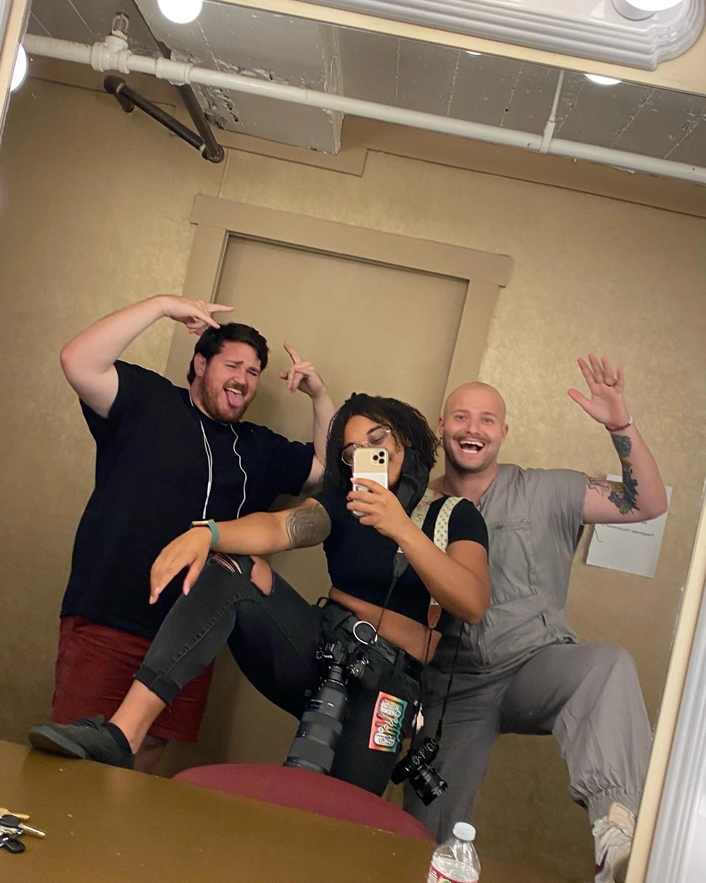 Three friends taking a mirror selfie inside a room, with one person sitting on a table and the other two standing behind. They are making playful gestures and smiling.