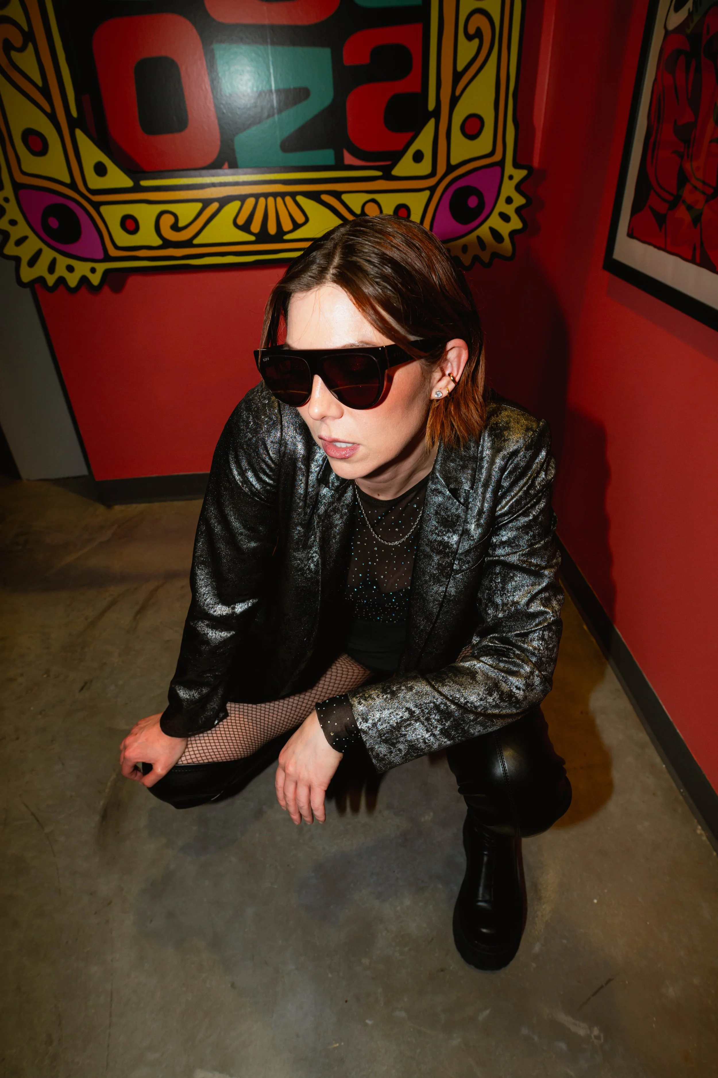 A woman wearing a shiny black blazer, fishnet stockings, and large black sunglasses crouching on the floor in front of a red and colorful illustrated wall.