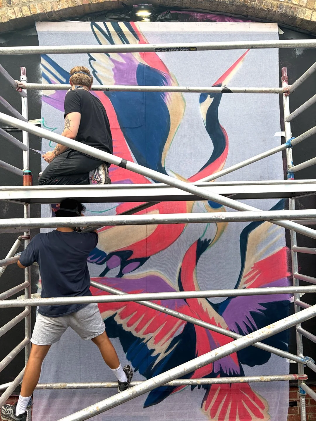 Two artists standing on a scaffold painting an image of birds on a large translucent canvas 
