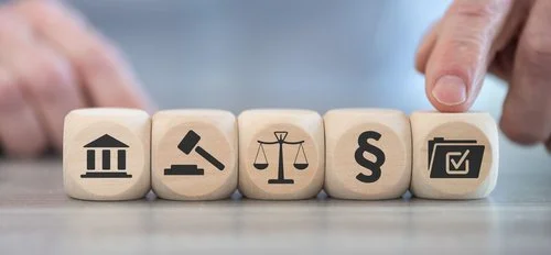 Five wooden blocks with legal and governmental symbols, including a courthouse, gavel, scales of justice, legal paragraph symbol, and a checklist, on a table.