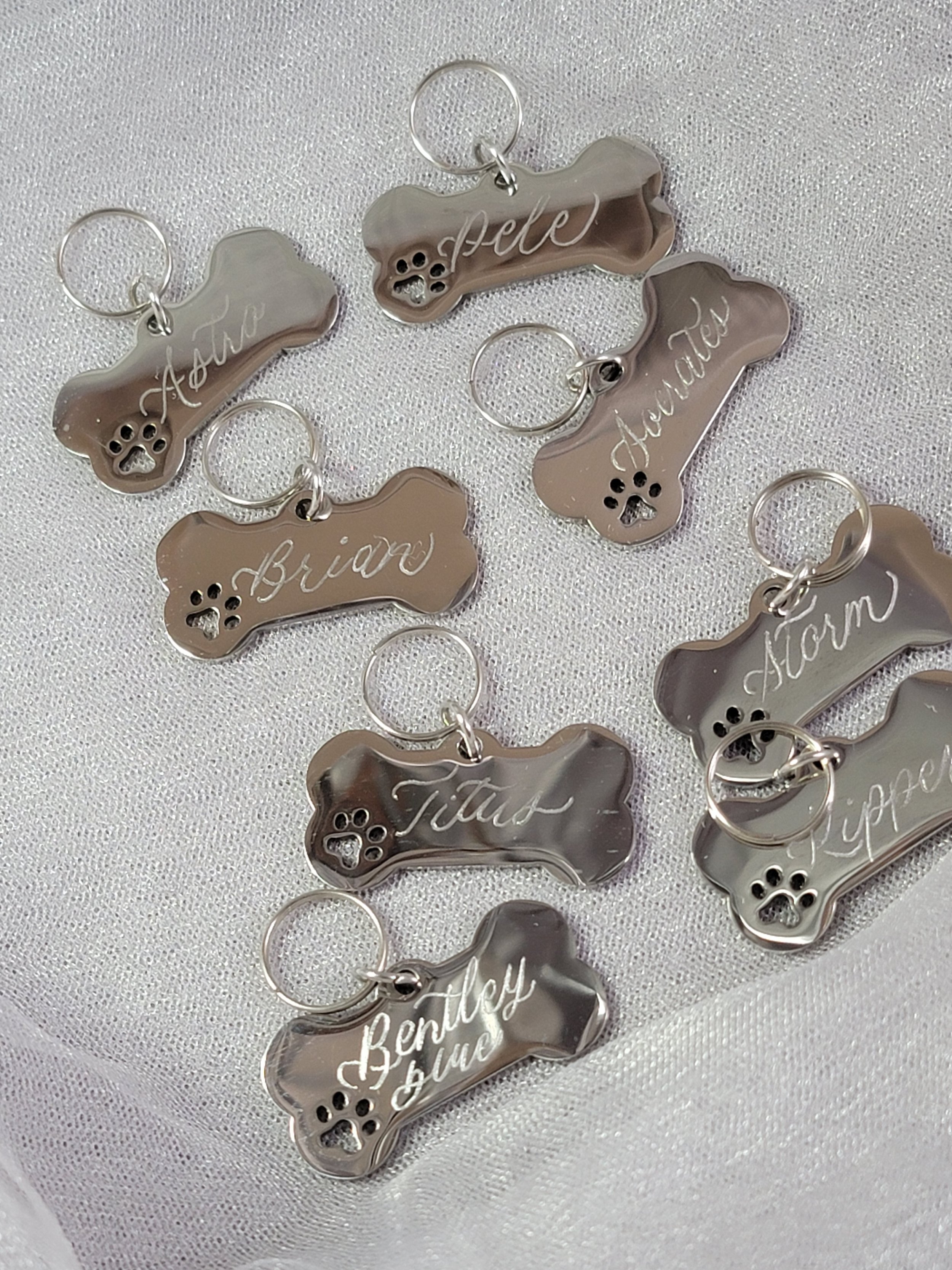 Multiple pet ID tags shaped like bones, engraved with the names Ash, Pete, and Benjamin, on a white textured surface.