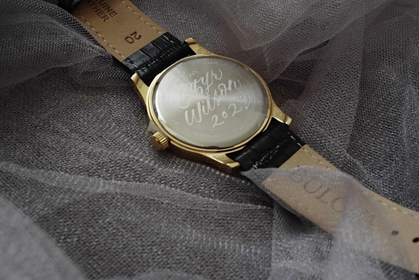 A wristwatch with a black leather strap and a gold case, displaying a message engraved on the back that reads 'Happy Will Power 2023'. The watch is resting on a soft, gray fabric surface.