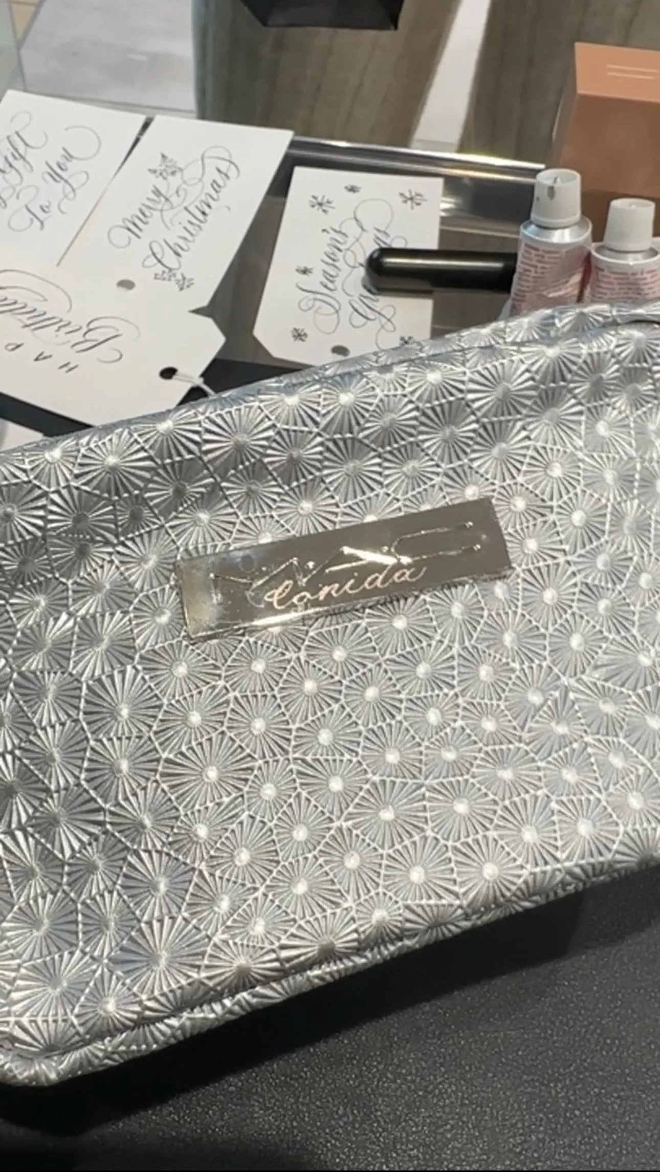A silver, patterned gift box with the name 'canda' on a metallic label. Behind the box, there are handwritten Holiday-themed cards and small bottles of white paint or lotion on a table.