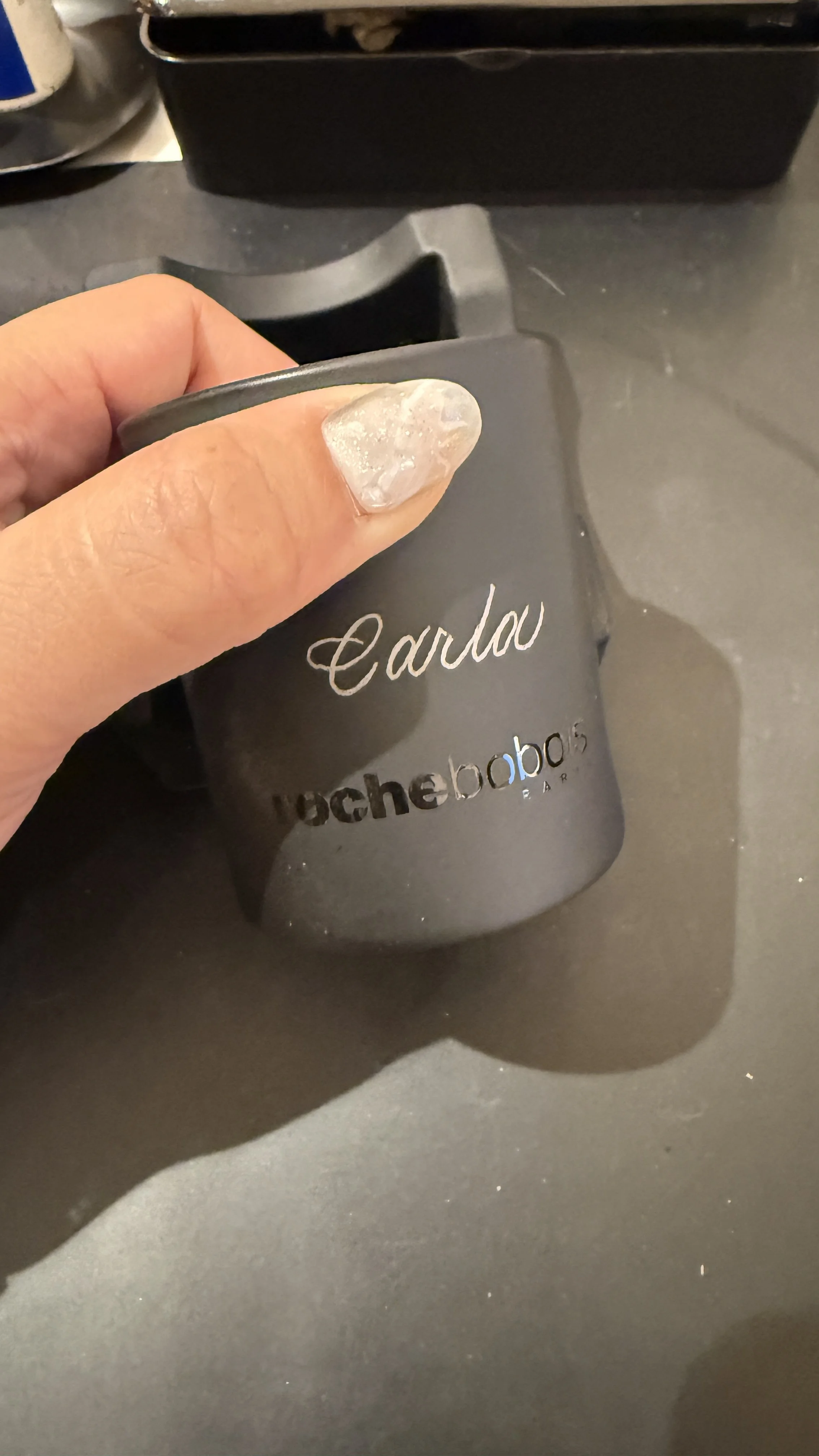 Close-up of a hand holding a small container with the text 'Carlo' and 'nocheboboc' on it, placed on a dark surface.