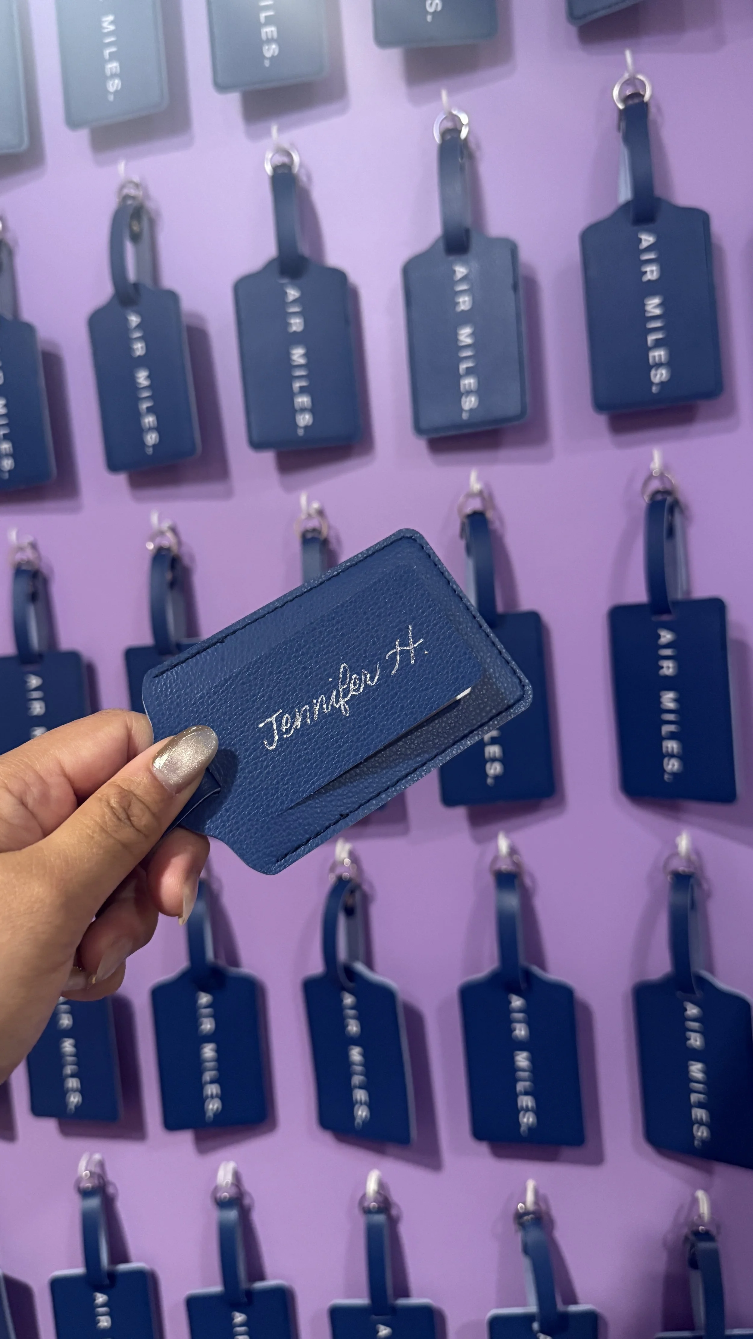 Hand holding a blue luggage tag with the name 'Jemalfer X.' in front of a wall display with multiple blue luggage tags labeled 'AIR MILES'. all tags are hanging in rows on a purple background.