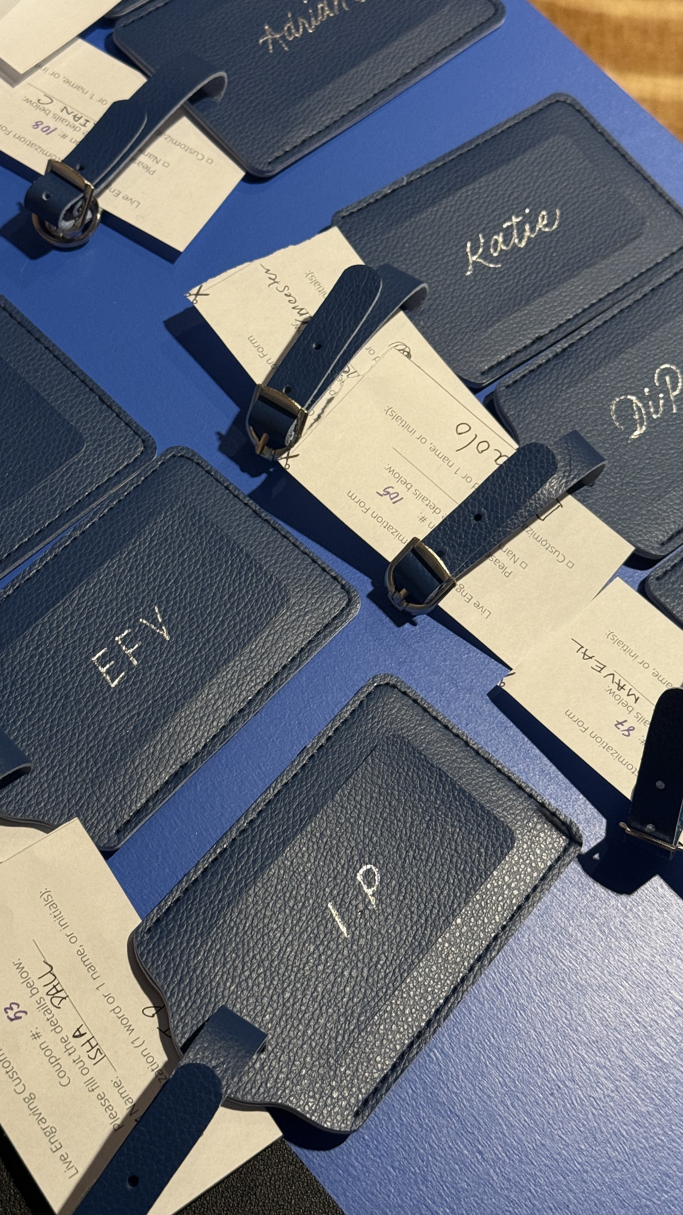 Multiple blue textured leather luggage tags with white embroidered names such as 'Katie', 'Diane', and 'Eileen', attached to blue surfaces, along with boarding passes and paper tags.