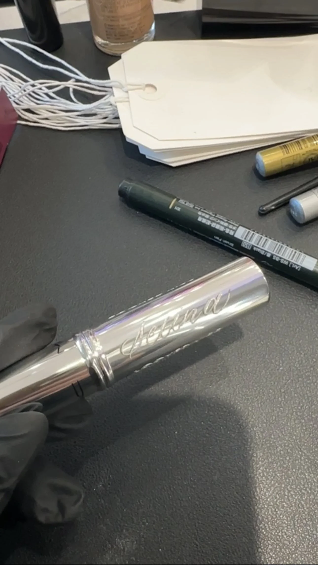 A black table surface with various items including white paper tags, a black marker, a golden marker, a white and gray marker, a jar, and a shiny metallic object with the word 'friends' engraved on it.