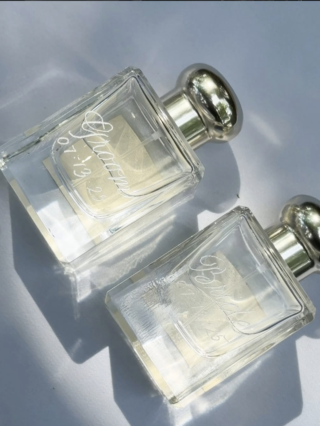 Two glass perfume bottles with silver caps on a light surface, casting shadows in sunlight.
