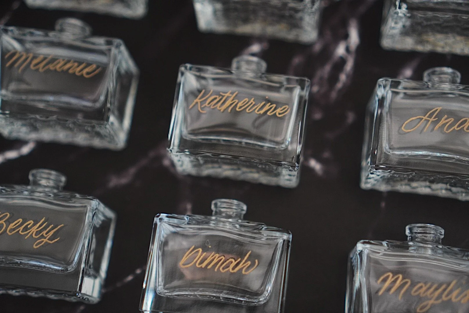 Close-up view of several glass perfume bottles with gold cursive writing of names, placed on a dark surface. The bottles are rectangular with rounded edges, each labeled with a different name such as Katherine, Becky, Dima, Anna, and Mayla.