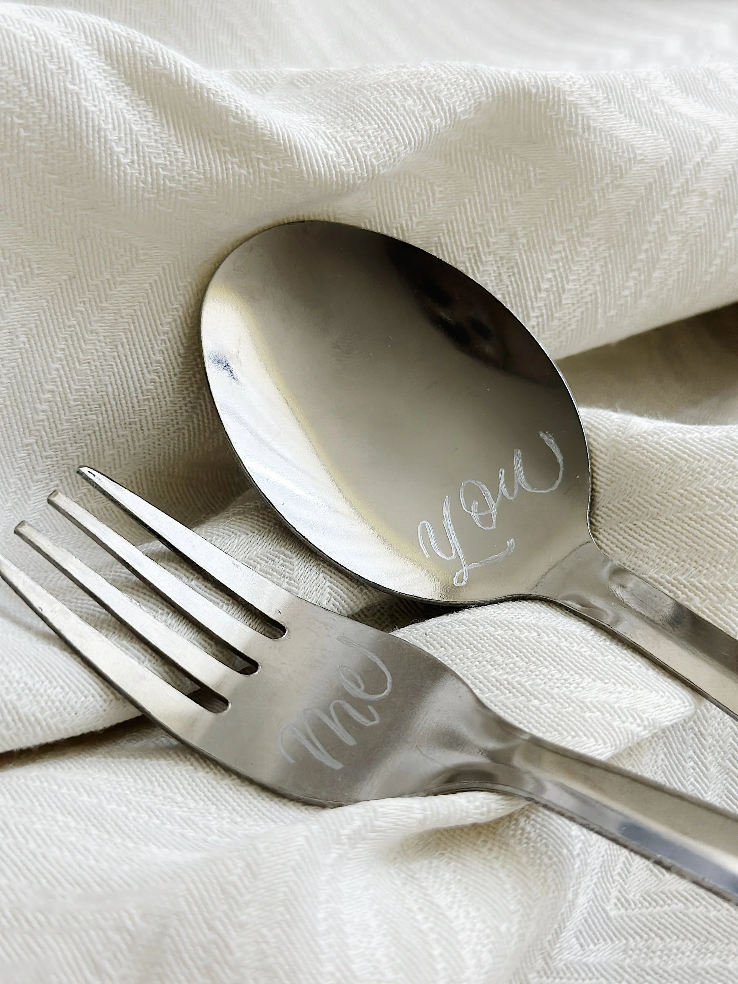 Close-up of a white napkin with a textured pattern, a silver spoon with the word 'love' engraved on it, and a silver fork resting on the napkin.