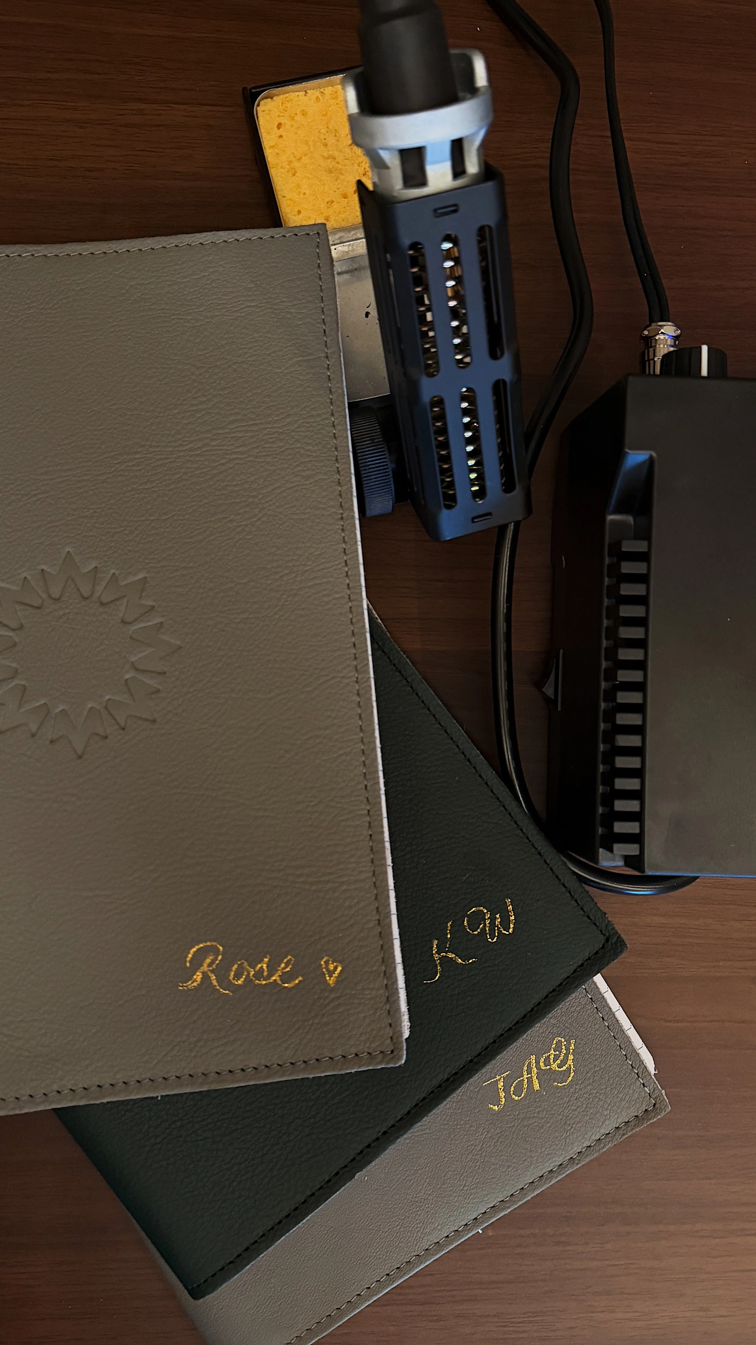 Three notebooks with gold-embossed names, 'Rose', 'Tacy', and 'Mary', stacked on a wooden surface, with a microphone, a sponge, and electronic equipment visible on the right.