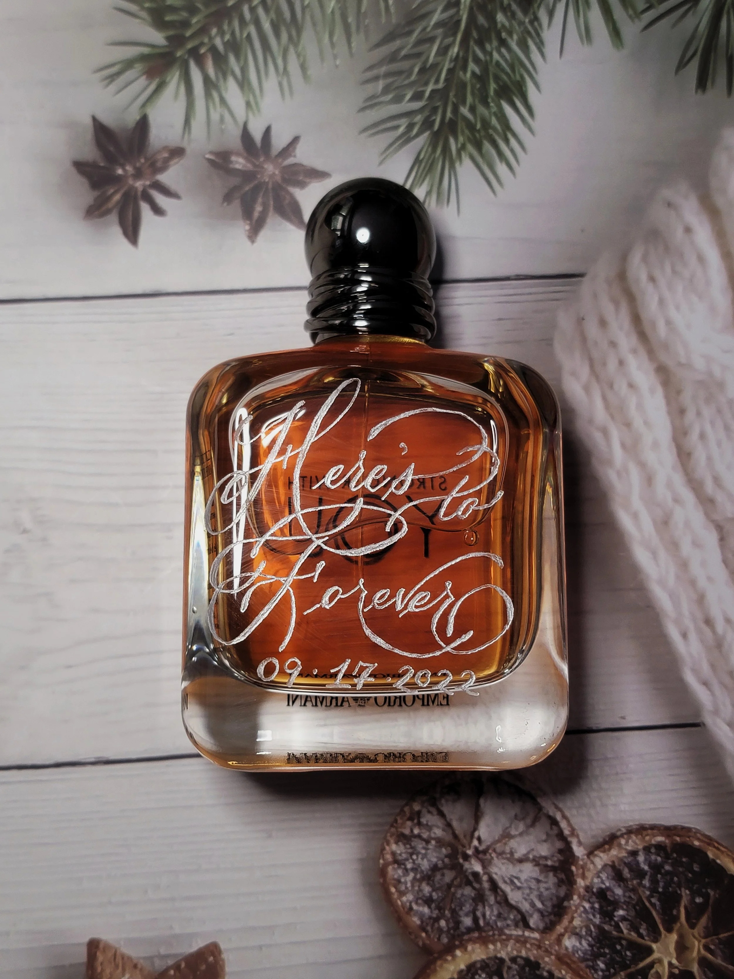 A glass perfume bottle with amber-colored liquid, engraved with the words "Here's to Forever" and a date, beneath a sprig of green pine, star anise, and dried orange slices on a white wooden surface.