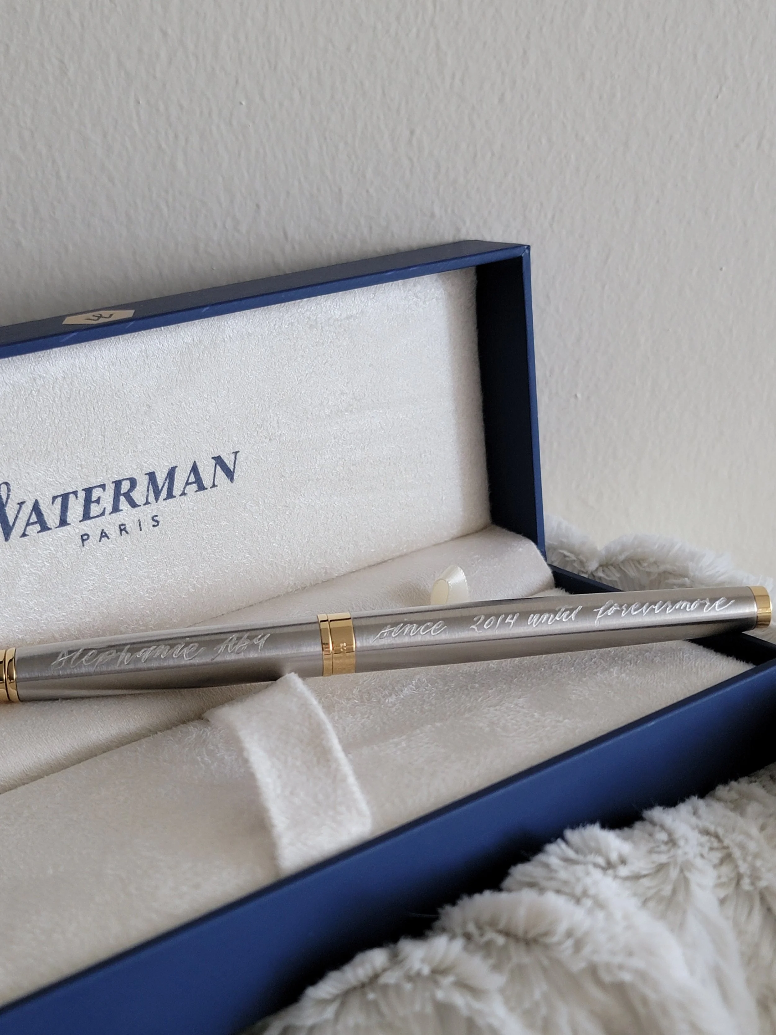 A silver pen with gold accents inside a blue jewelry box, which has a white satin lining and the words 'since 2014 under forever' engraved on it, resting on a plush white surface.