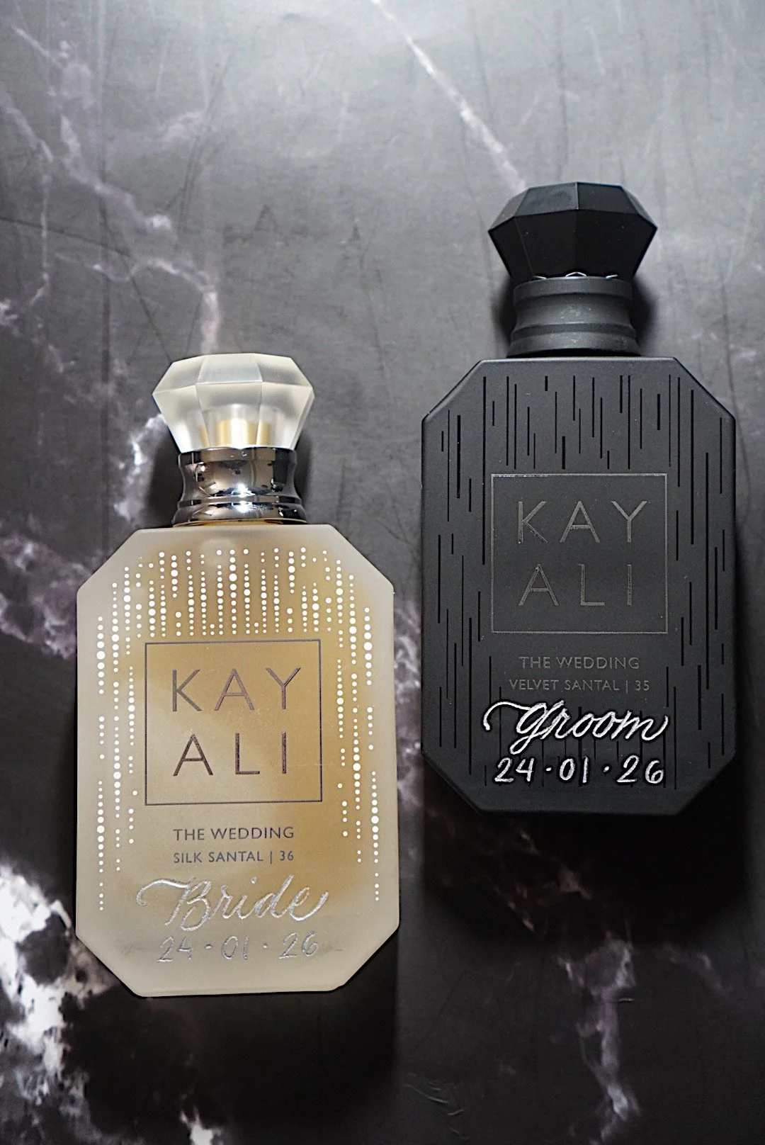 Two perfume bottles, one cream-colored labeled 'Bride' and the other black labeled 'Groom', on a dark marble surface.