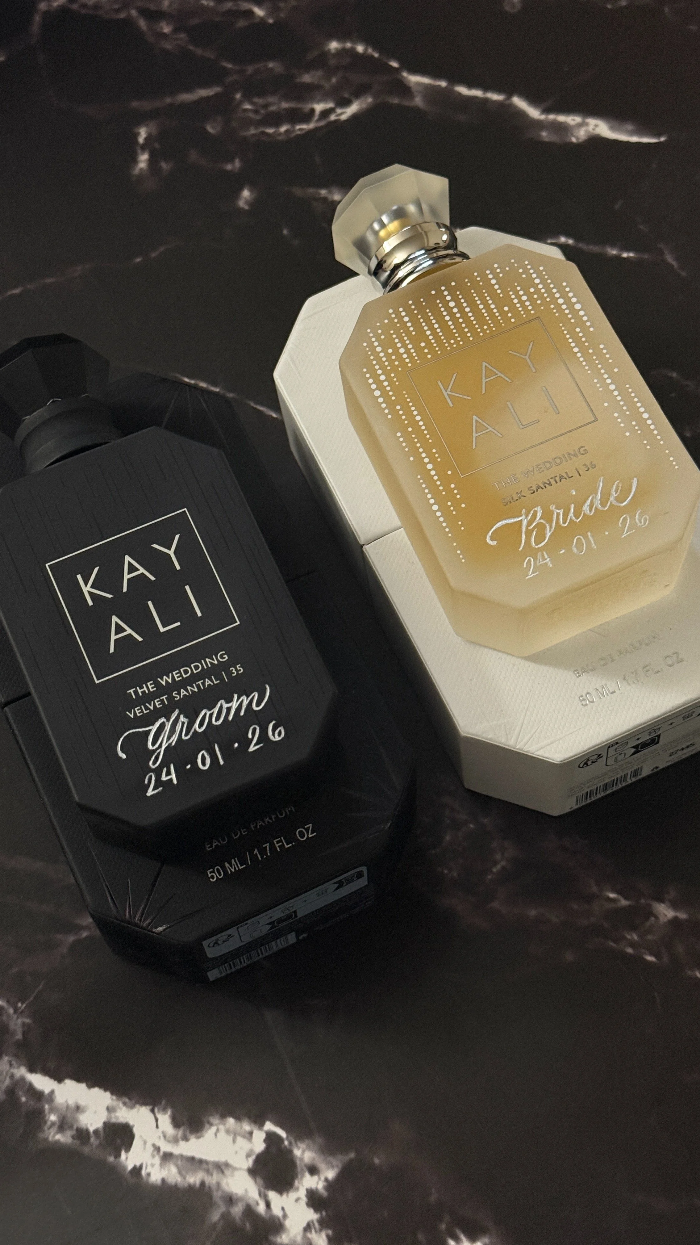 Two bottles of Kay Ali perfume on a dark marble surface, one labeled 'Groom' and the other labeled 'Bride', with details about wedding date and scent type.