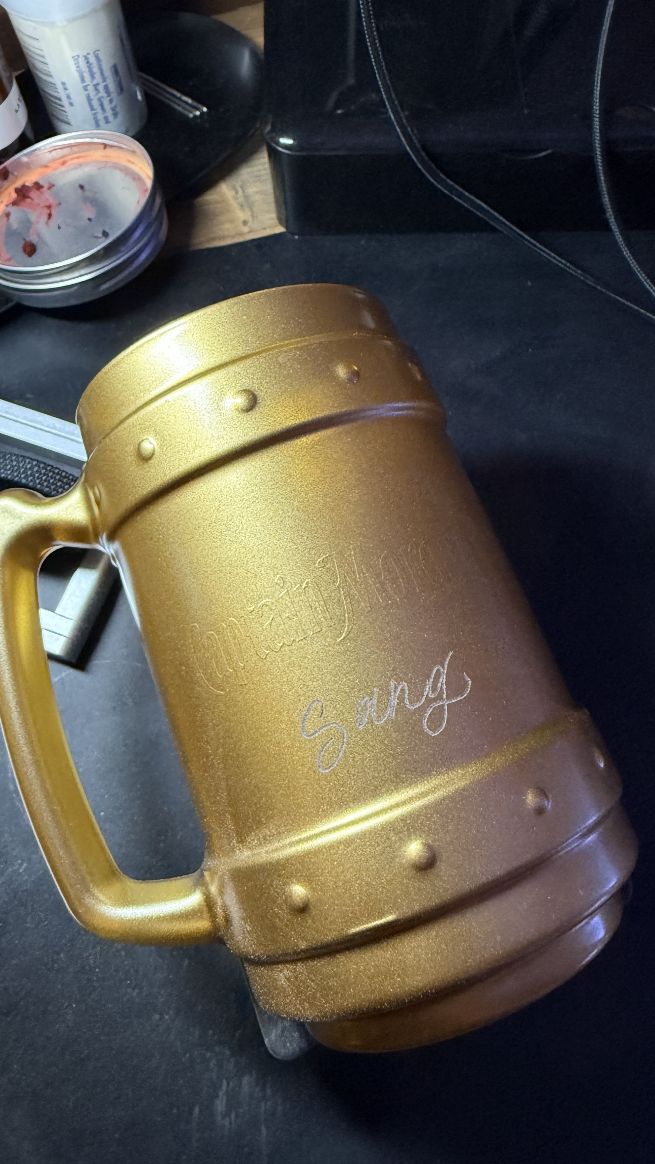 A large, gold-colored mug with the word "Sunday" engraved on it, set on a dark surface with some objects and a phone in the background.