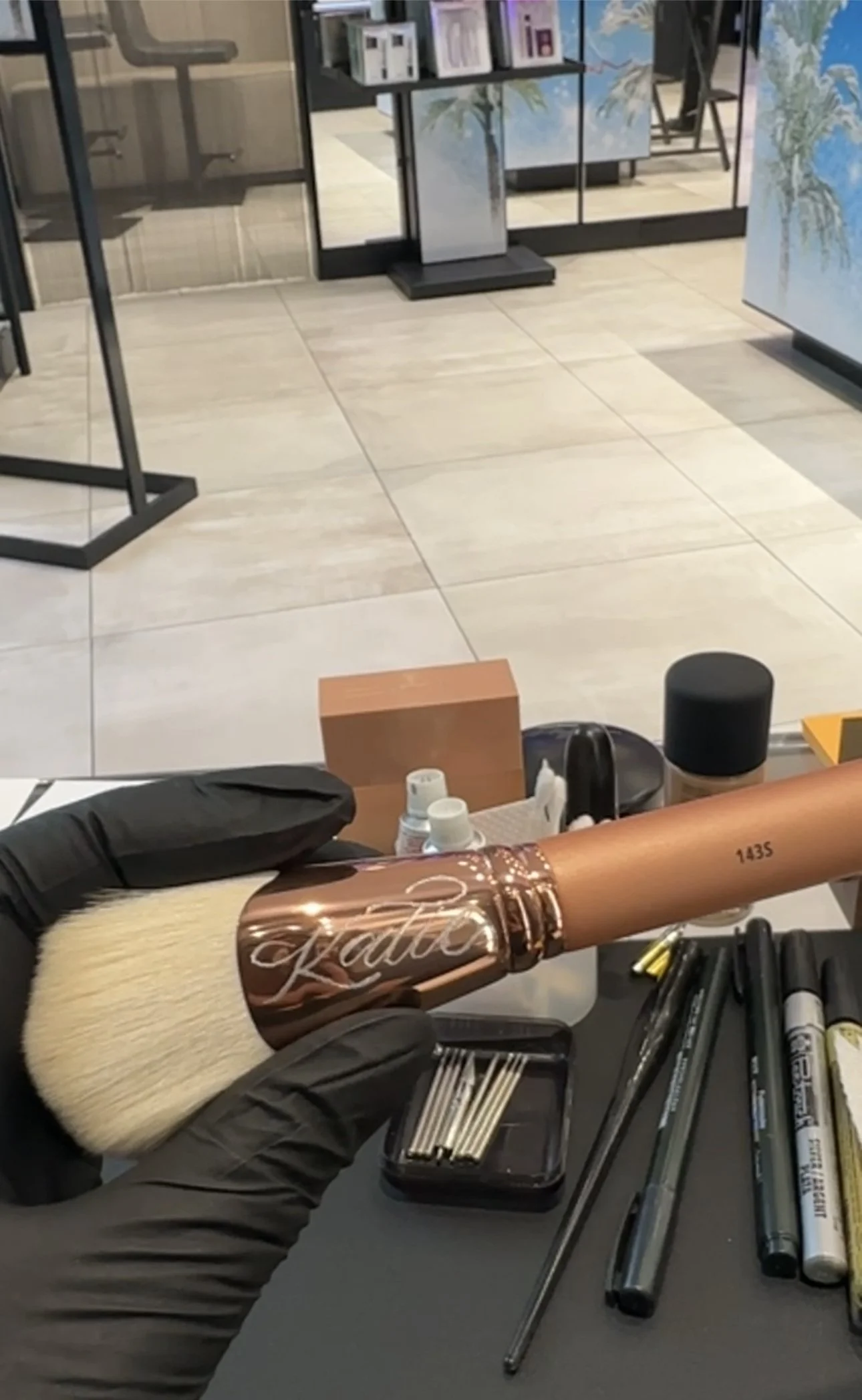 A person wearing black gloves holding a makeup brush with a rose gold handle, engraved with the name 'Kaitlin,' in a makeup station with various makeup tools and supplies on a black surface. In the background, there is a tiled floor and glass storefr