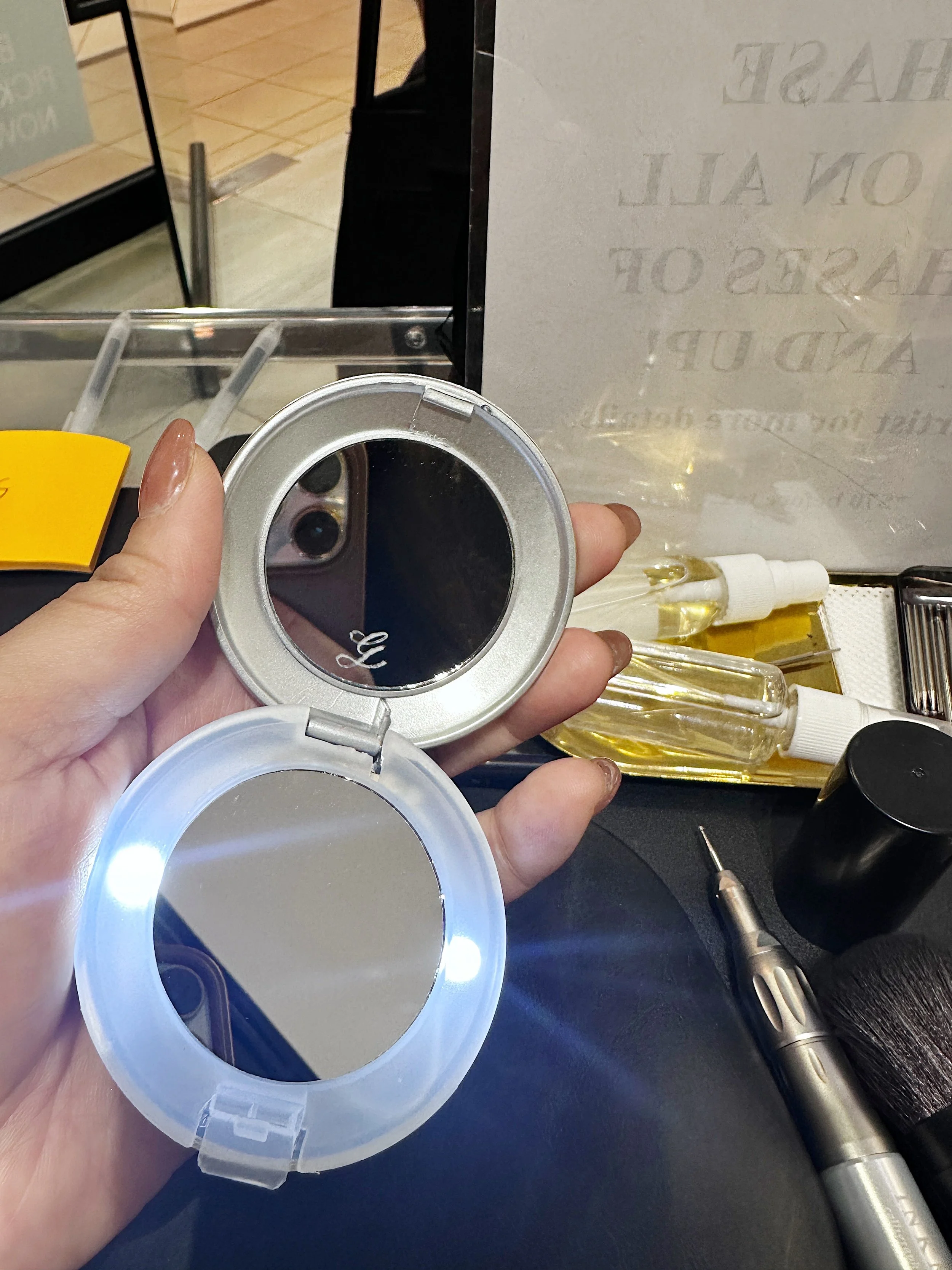 A person's hand holding a metallic round container with a reflective interior, surrounded by dental or medical tools, bottles, and a tissue on a dark surface.