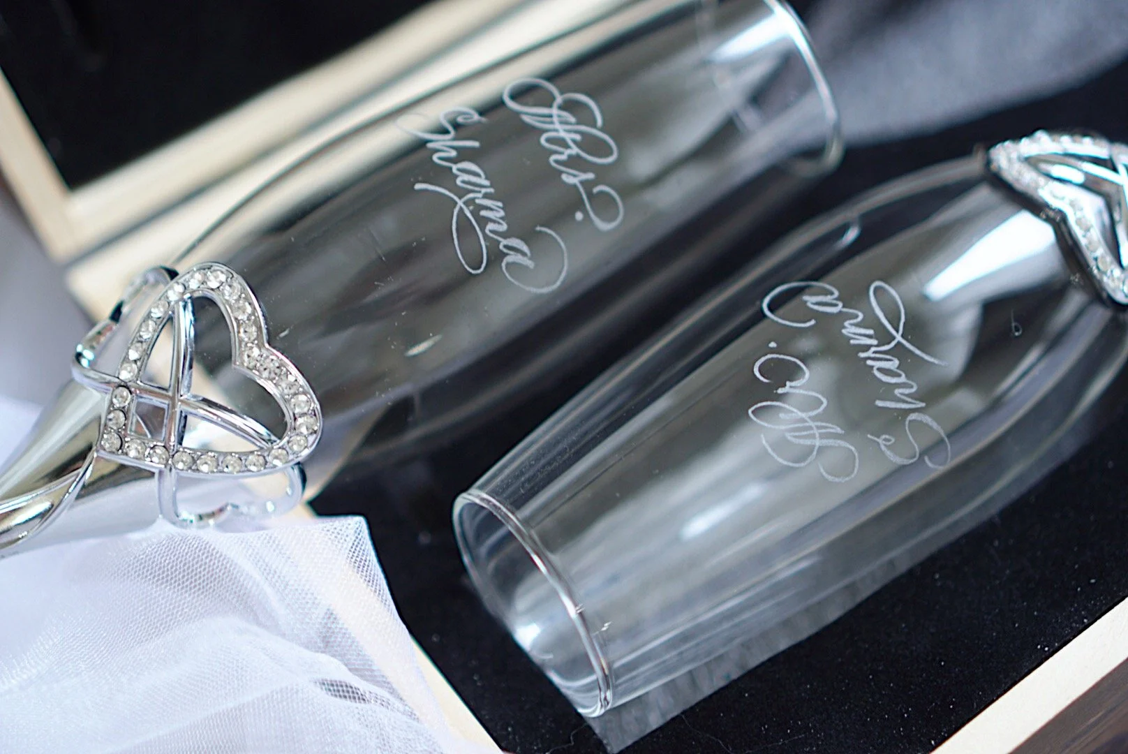 Close-up of two clear glass champagne flutes with white cursive writing on them, resting on a black surface. One flute has a silver heart-shaped decoration encrusted with rhinestones attached to it.