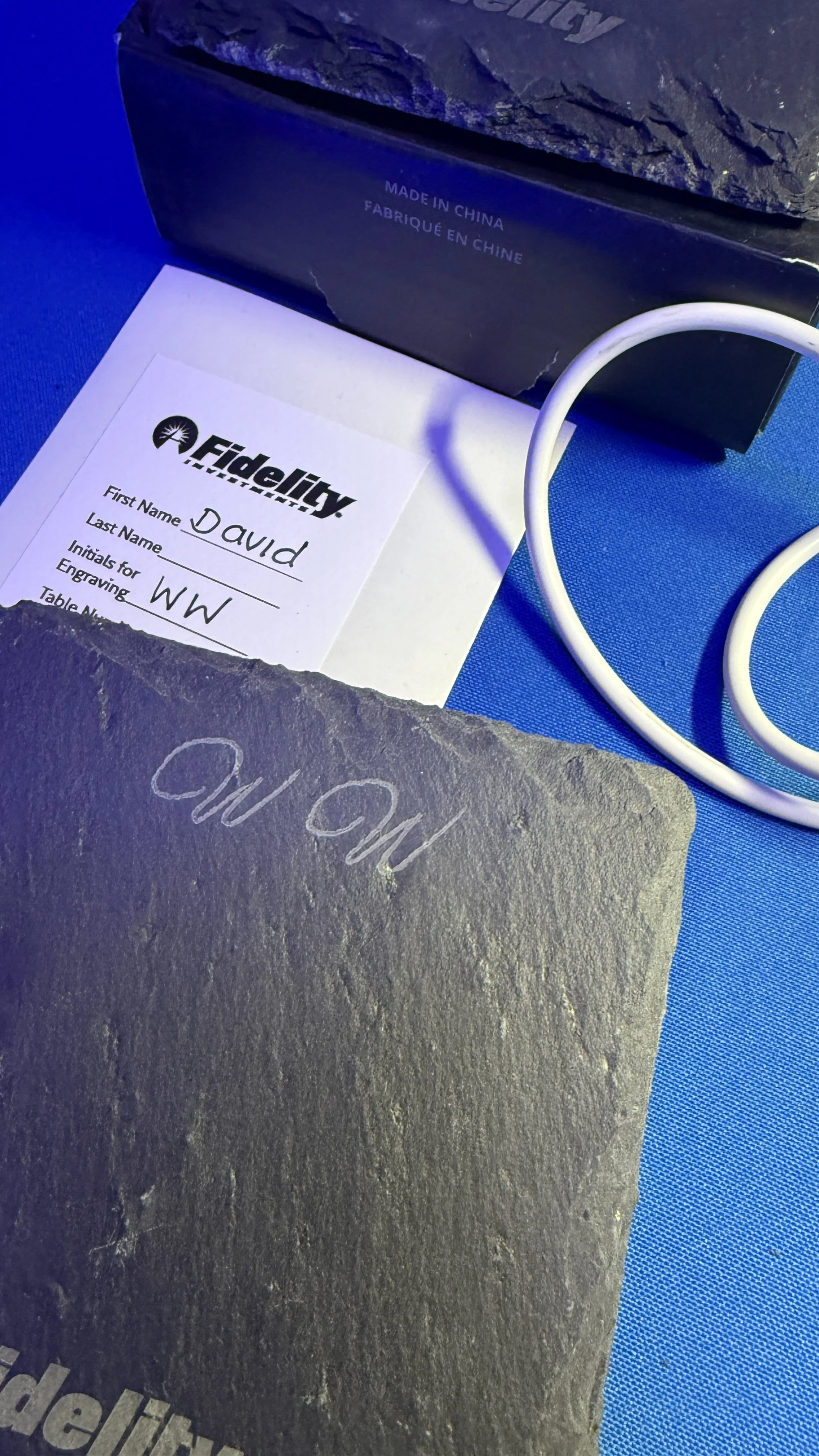 Close-up of a slate engraved with 'MO MO', a Fidelity Rewards program card with handwritten name 'David', initials 'WW', and a partially visible black box with 'Made in China' text. A white cable is also present.