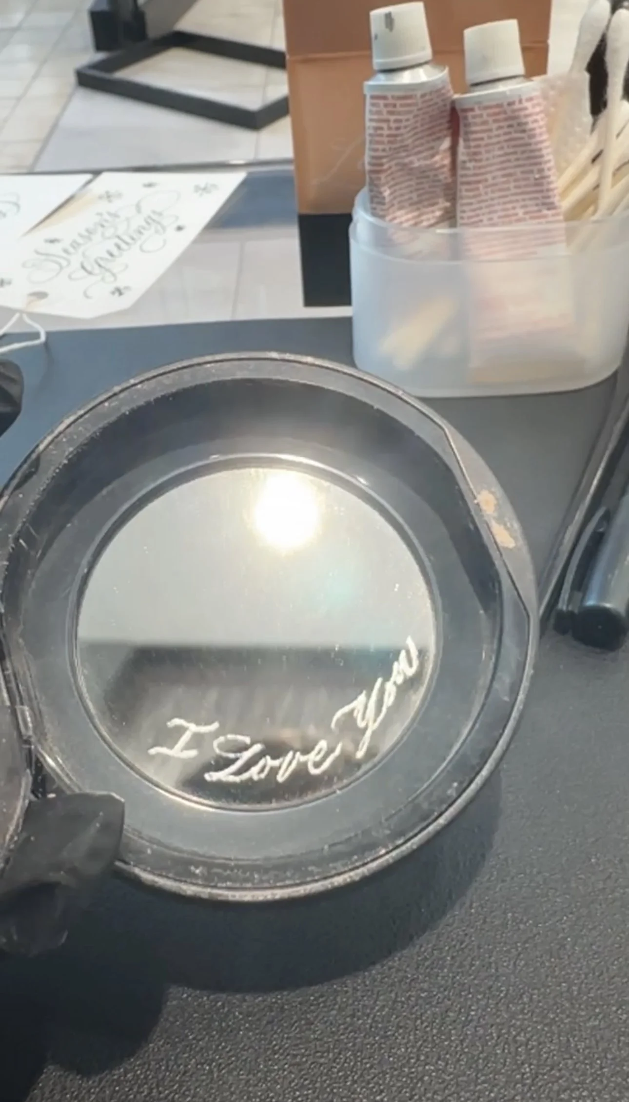 A small round mirror with the words 'I love you' written on it, placed on a dark table. In the background, there are various items including a container with spray cans and other objects.